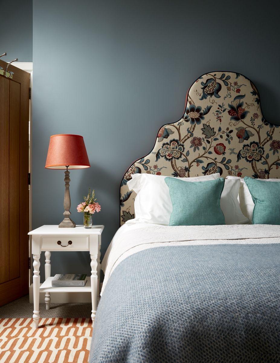 A bedroom with a floral headboard, teal cushions and white bedding against blue grey walls, with a lamp on a white bedside table.