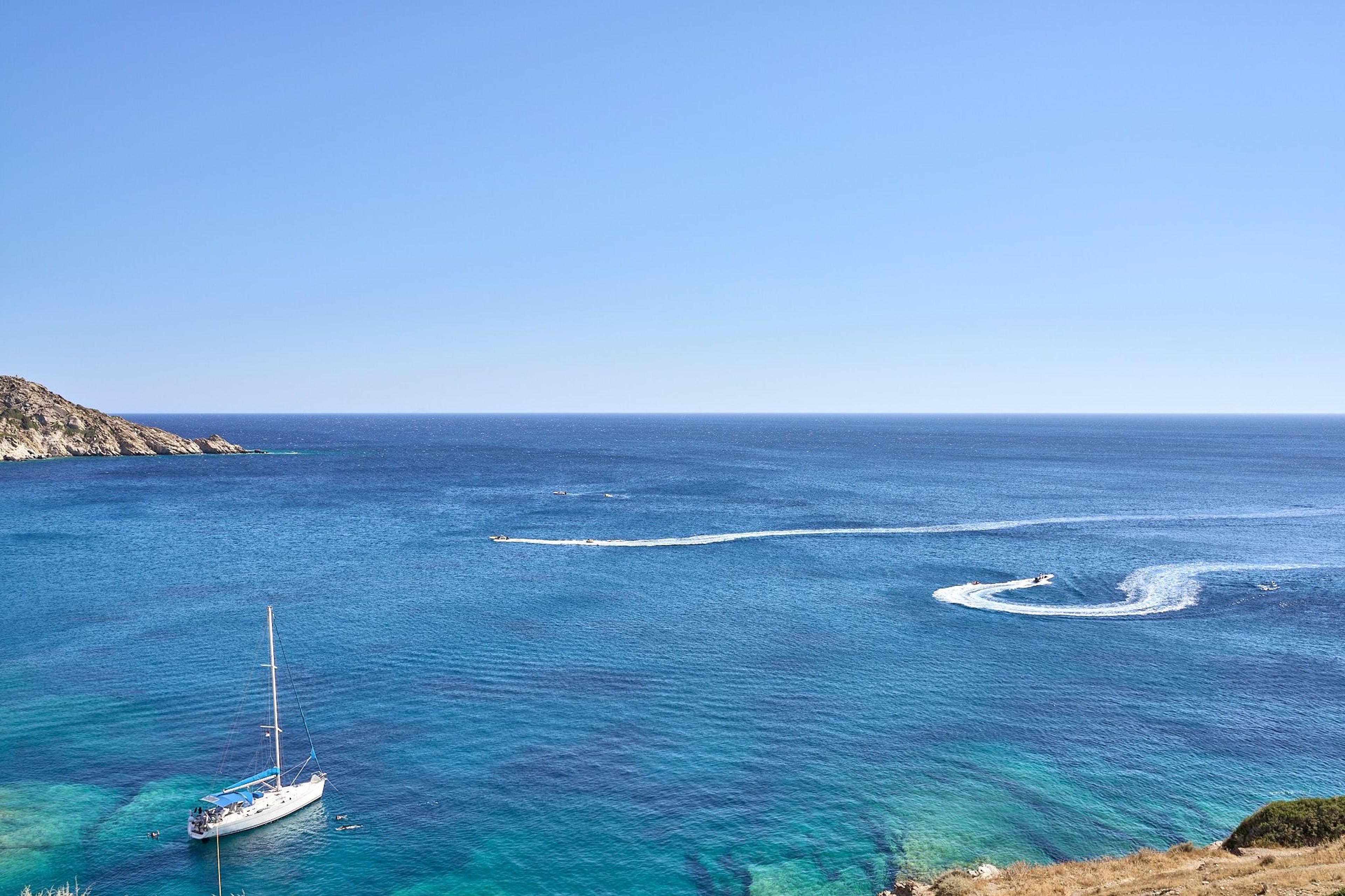 Open sea seen from above a headland, with a sailing boat at the lower left and the wake of a speedboat curving across the water.