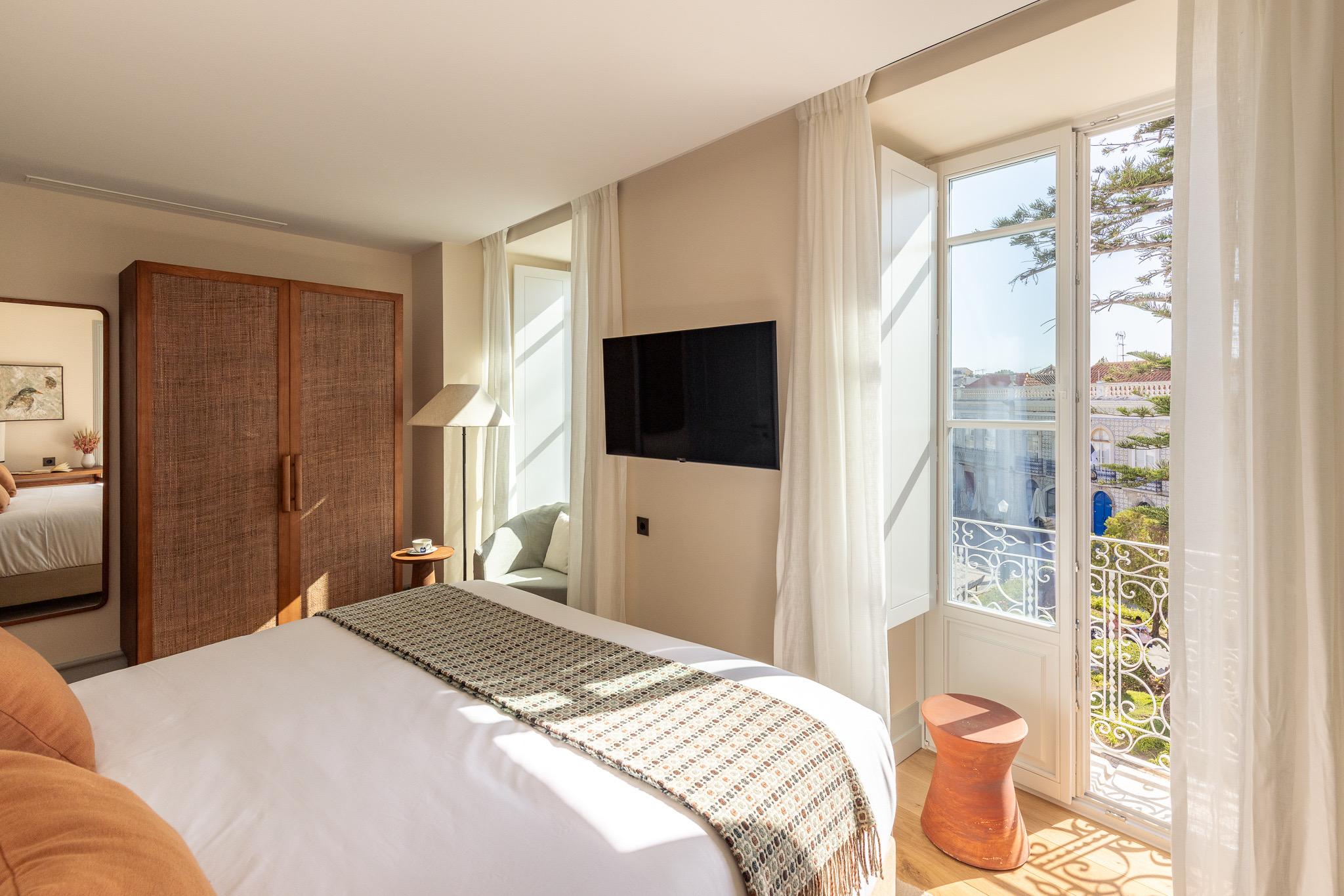 A hotel bedroom with a white bed and woven headboard, a wardrobe, and tall windows open to a wrought iron balcony overlooking a tree-lined square.