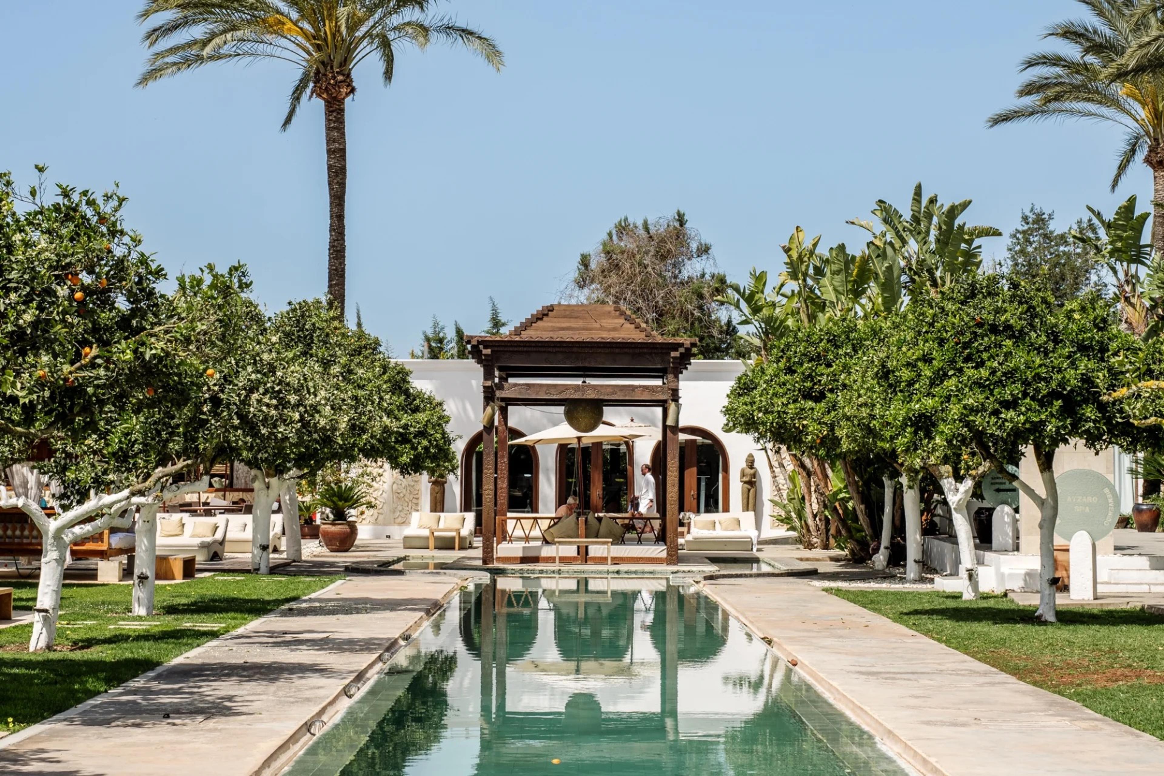 A long reflecting pool running between lawns and citrus trees to a carved wooden gazebo in front of the white main building.
