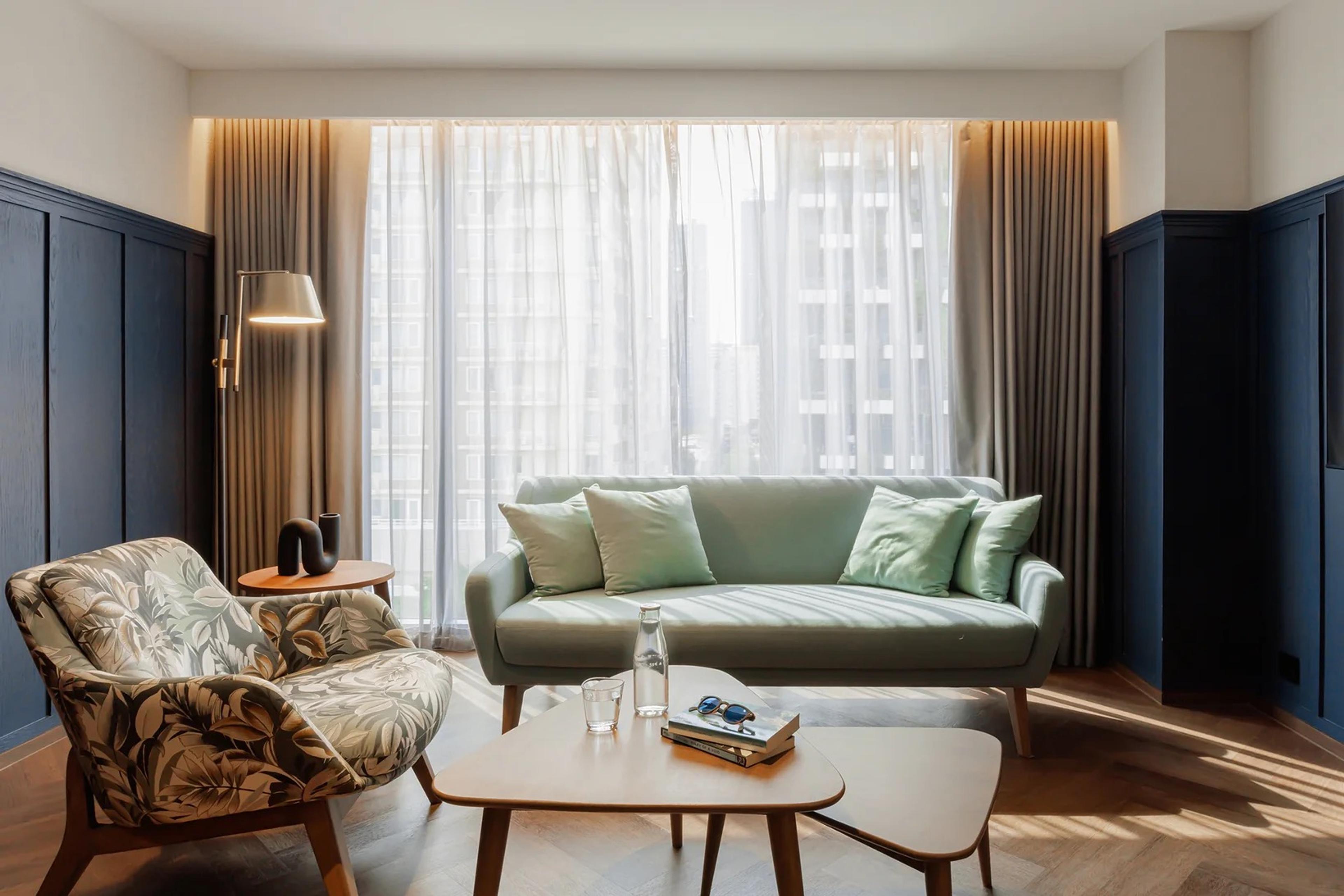 A living room with a pale green sofa, a floral print armchair and a low coffee table, lit by sheer curtains in front of a window.