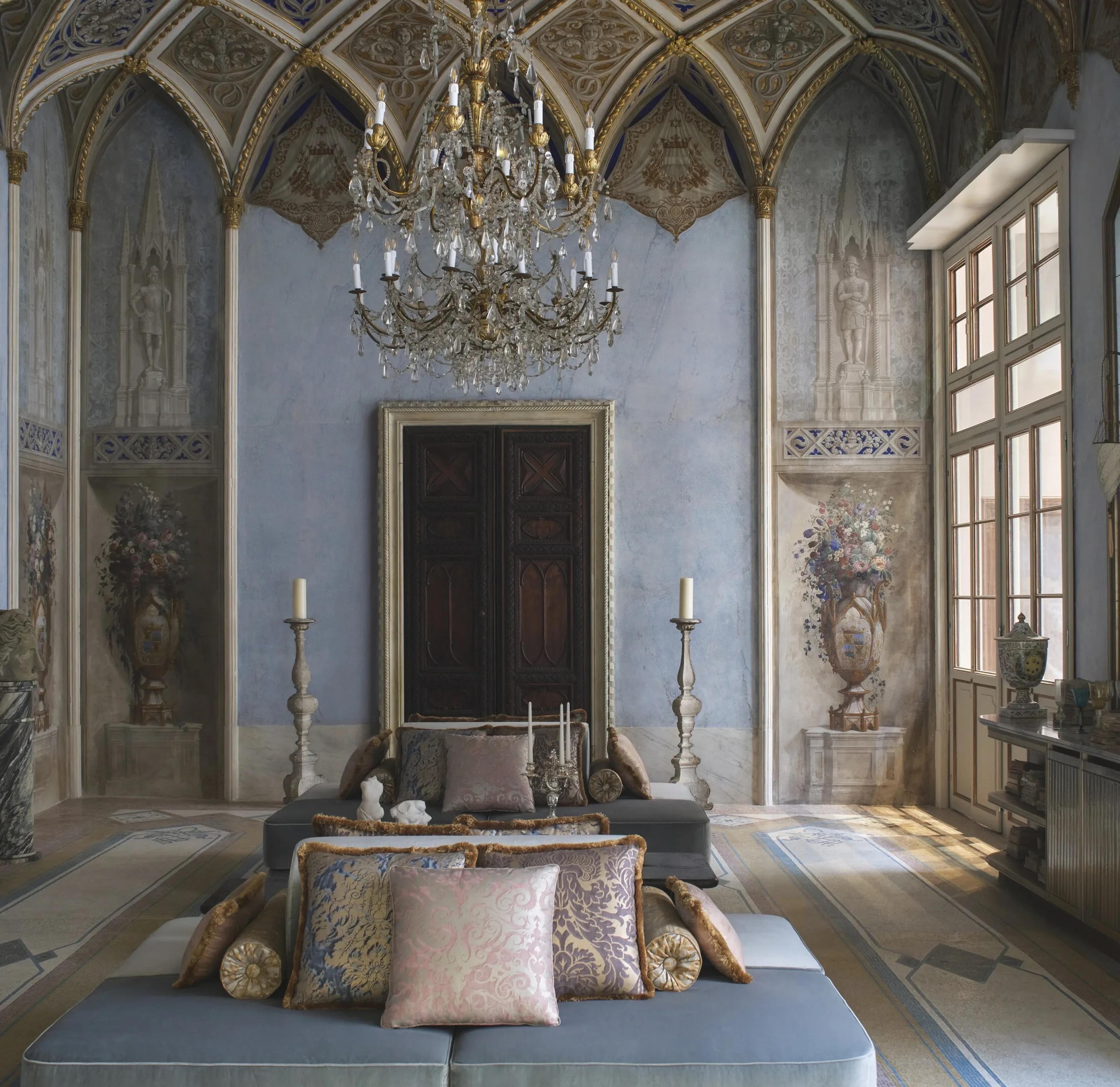 A frescoed blue hall with Gothic arched windows, a crystal chandelier, and two pale blue upholstered benches heaped with patterned cushions