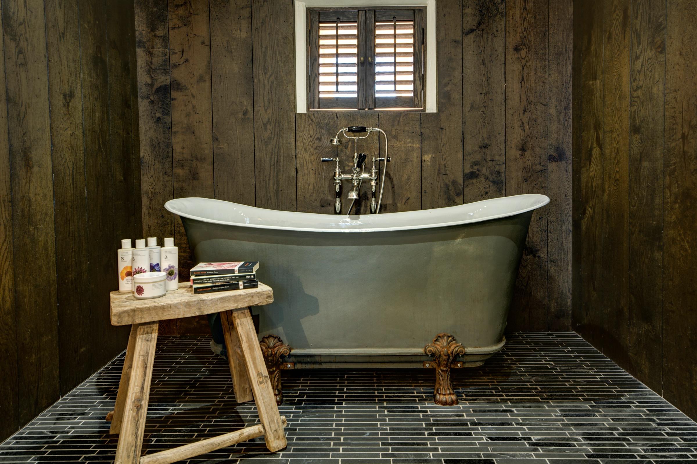 A freestanding bathtub with an old-fashioned tap on clawed feet, in a dark wood-panelled bathroom with a shuttered window and toiletries on a wooden stool.