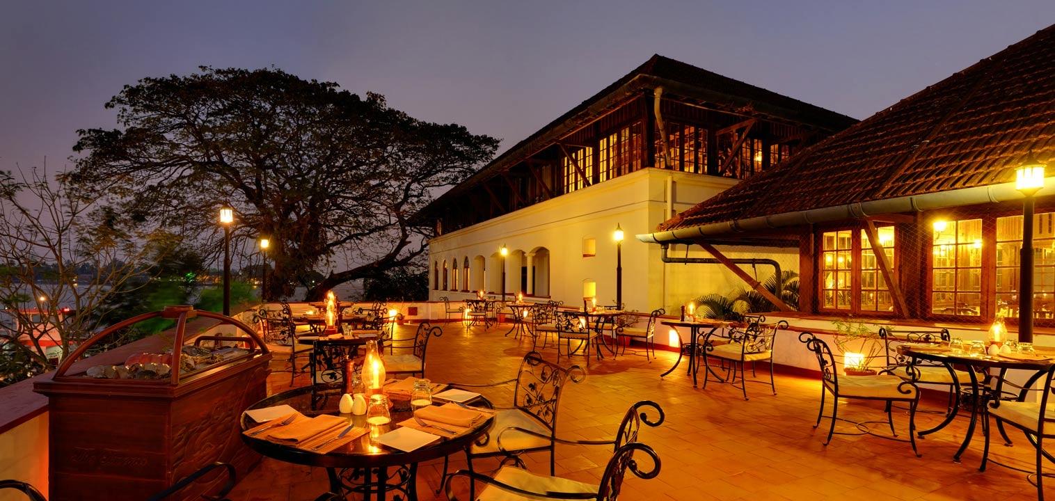 Wrought-iron tables laid for dinner on a stone terrace at dusk, with the hotel's lit facade and a large tree behind.