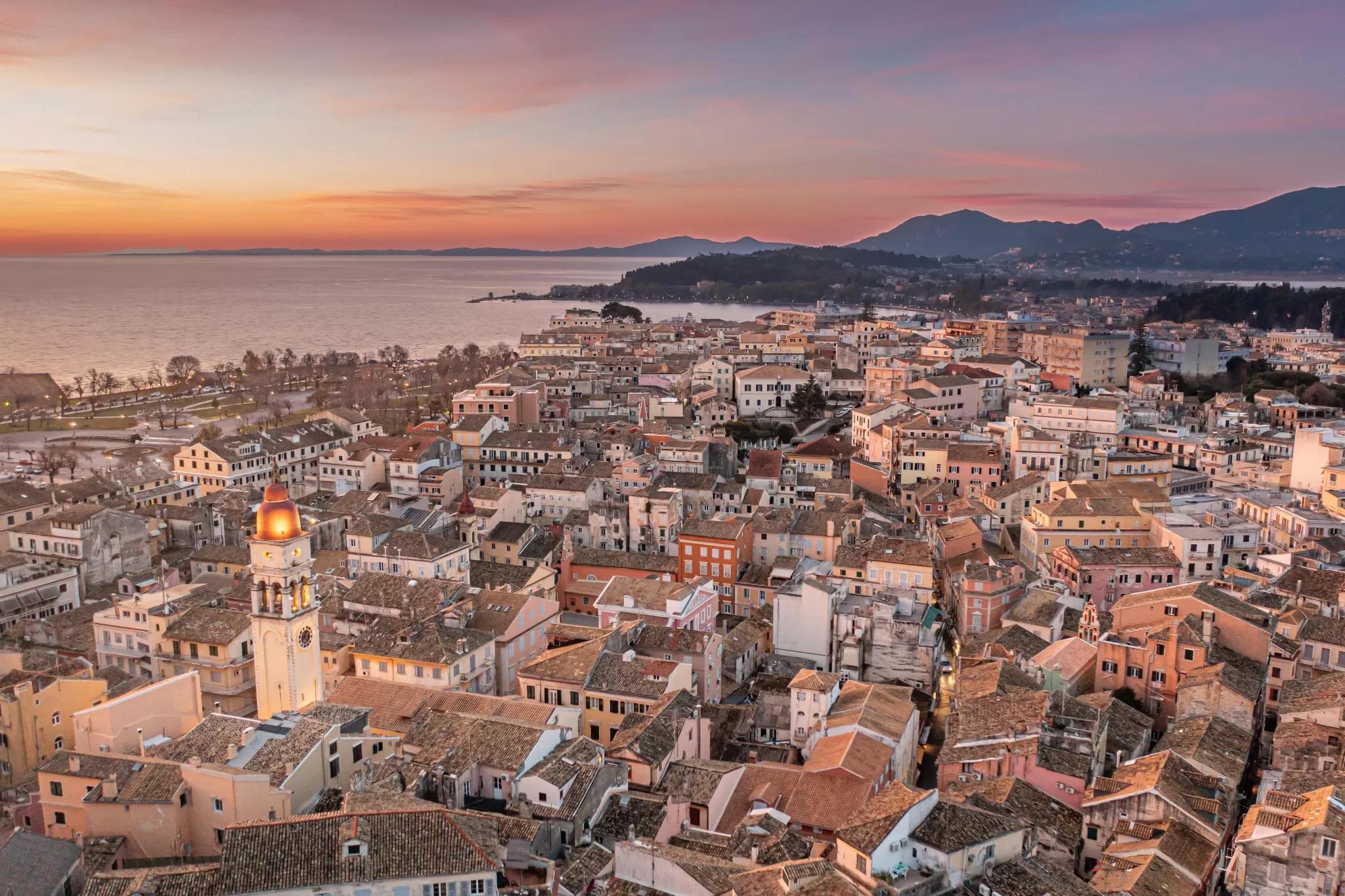 An aerial view at sunset of Corfu Town's terracotta rooftops and a domed bell tower, with the sea and hills beyond.