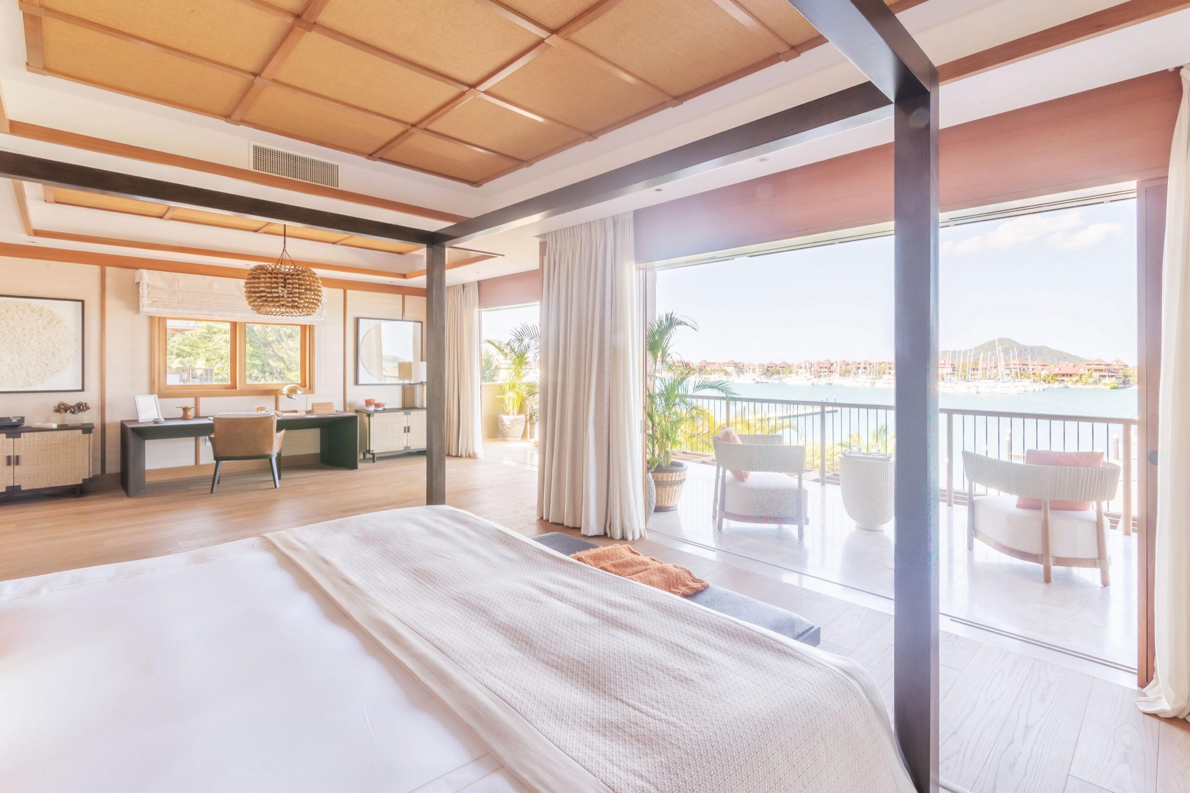 A bedroom with a white bed facing an open terrace, with armchairs and a view over a marina of moored boats.