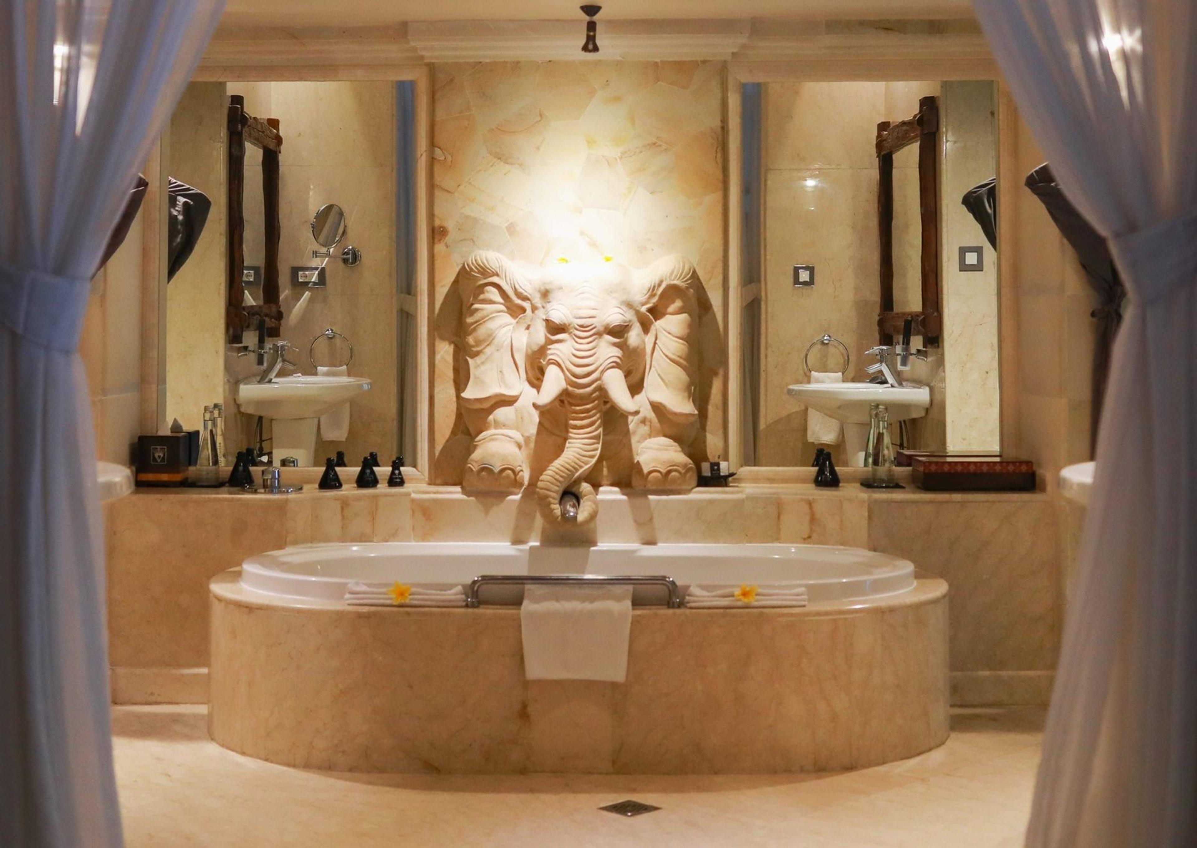 A round marble bathtub sits beneath a carved stone elephant head fountain, flanked by two sinks and mirrors framed by sheer curtains.