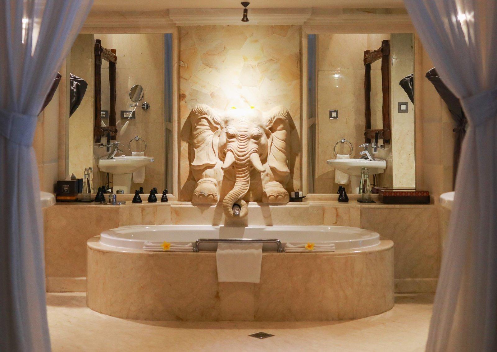 A round marble bathtub sits beneath a carved stone elephant head fountain, flanked by two sinks and mirrors framed by sheer curtains.