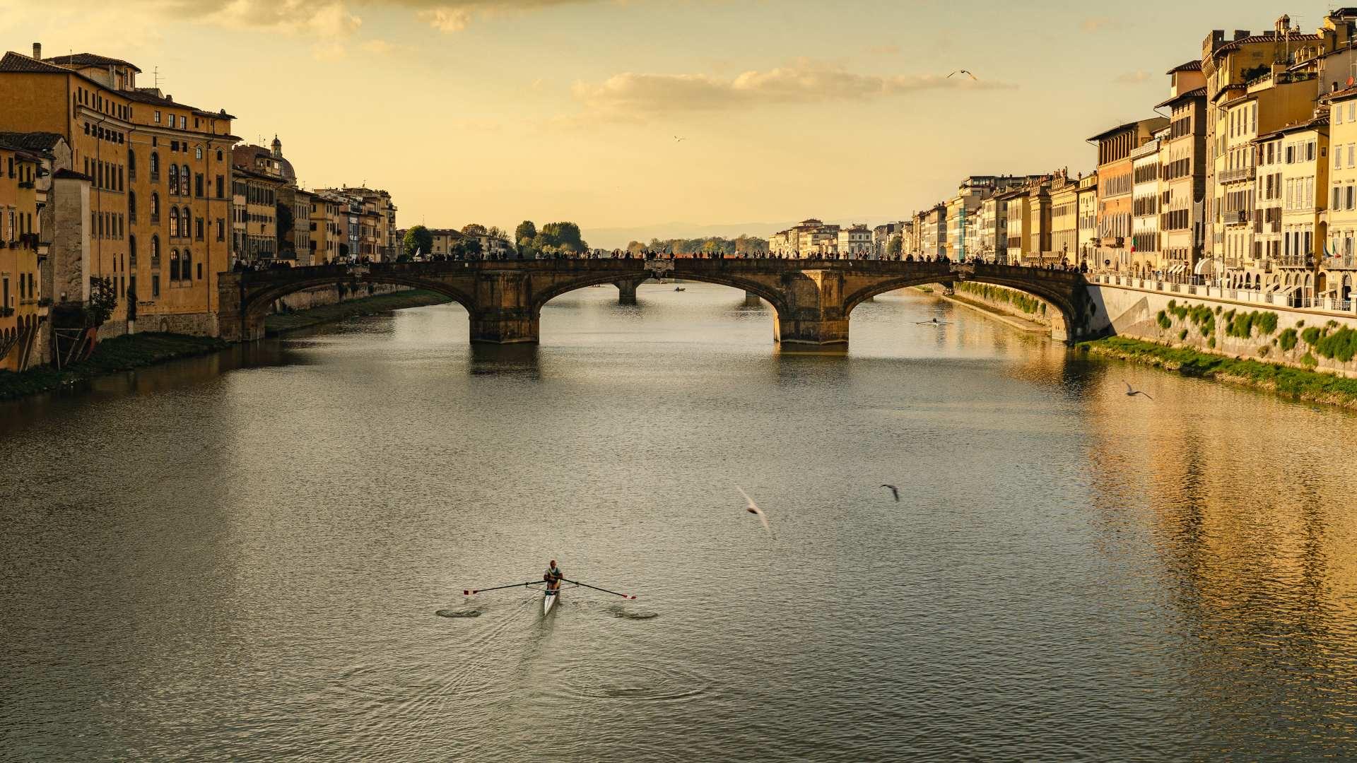 A view along the Arno at golden hour, a stone bridge crossing the river, a single rower in a scull on the water, and gulls in flight.