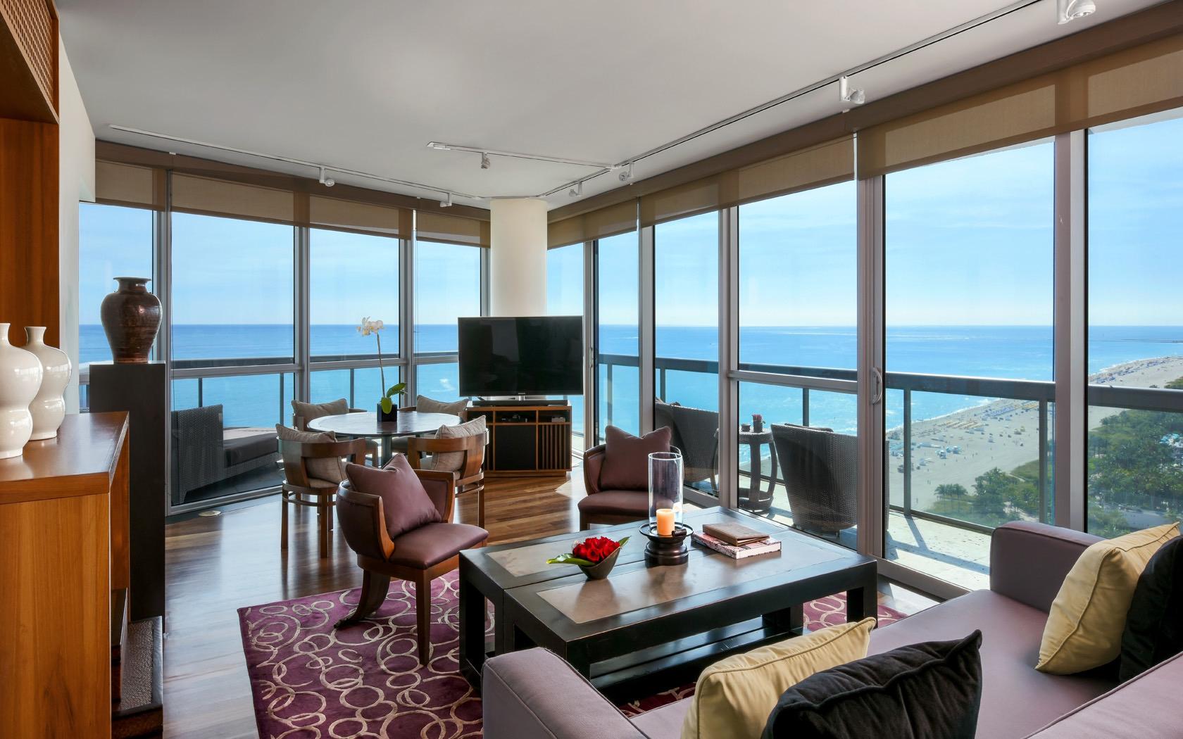 A high-rise suite's living and dining area with floor-to-ceiling windows overlooking the ocean and beach below.