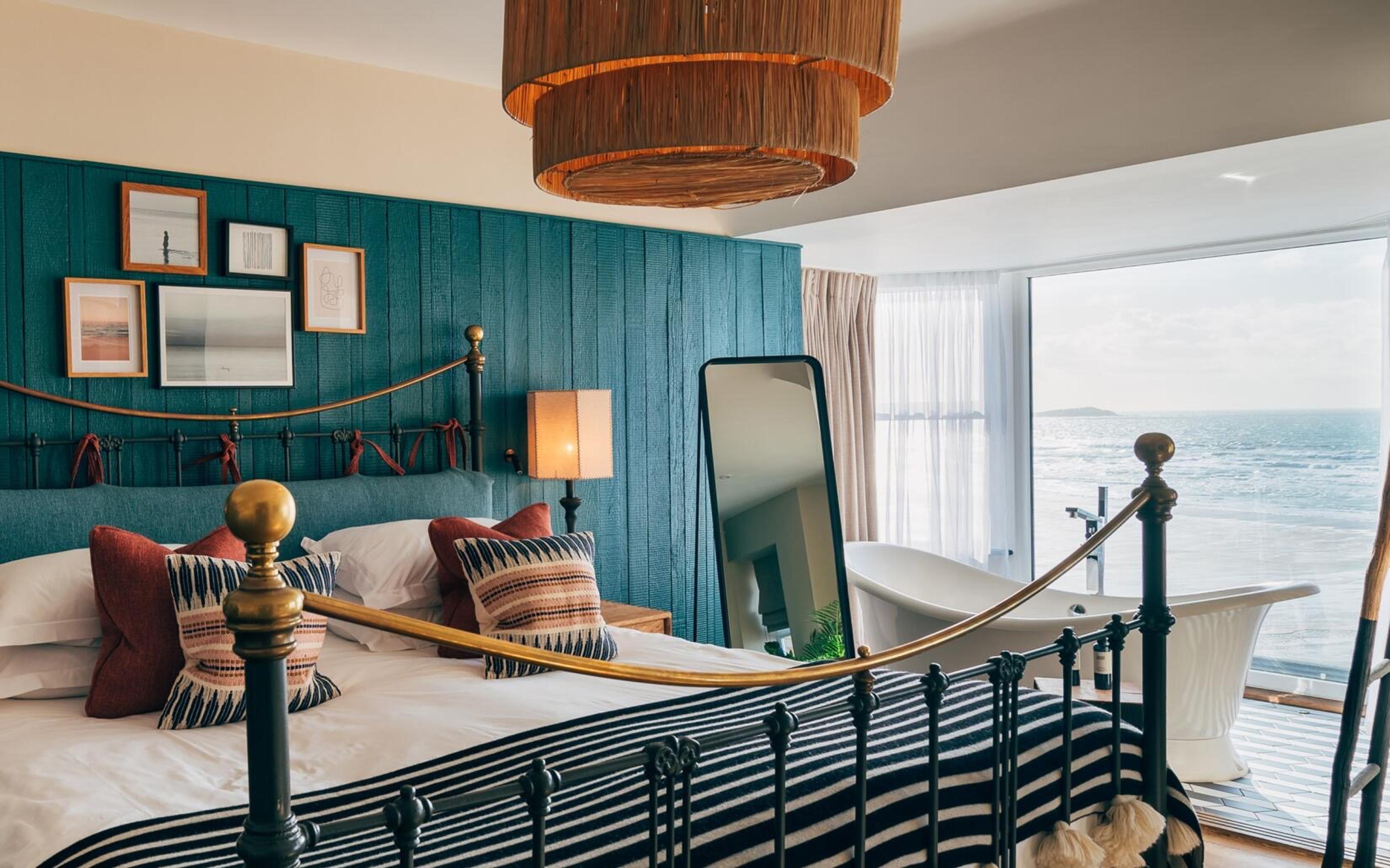 A bedroom with teal wood panelling, an iron bedstead with striped bedding, and a freestanding bath beside a window onto the sea.