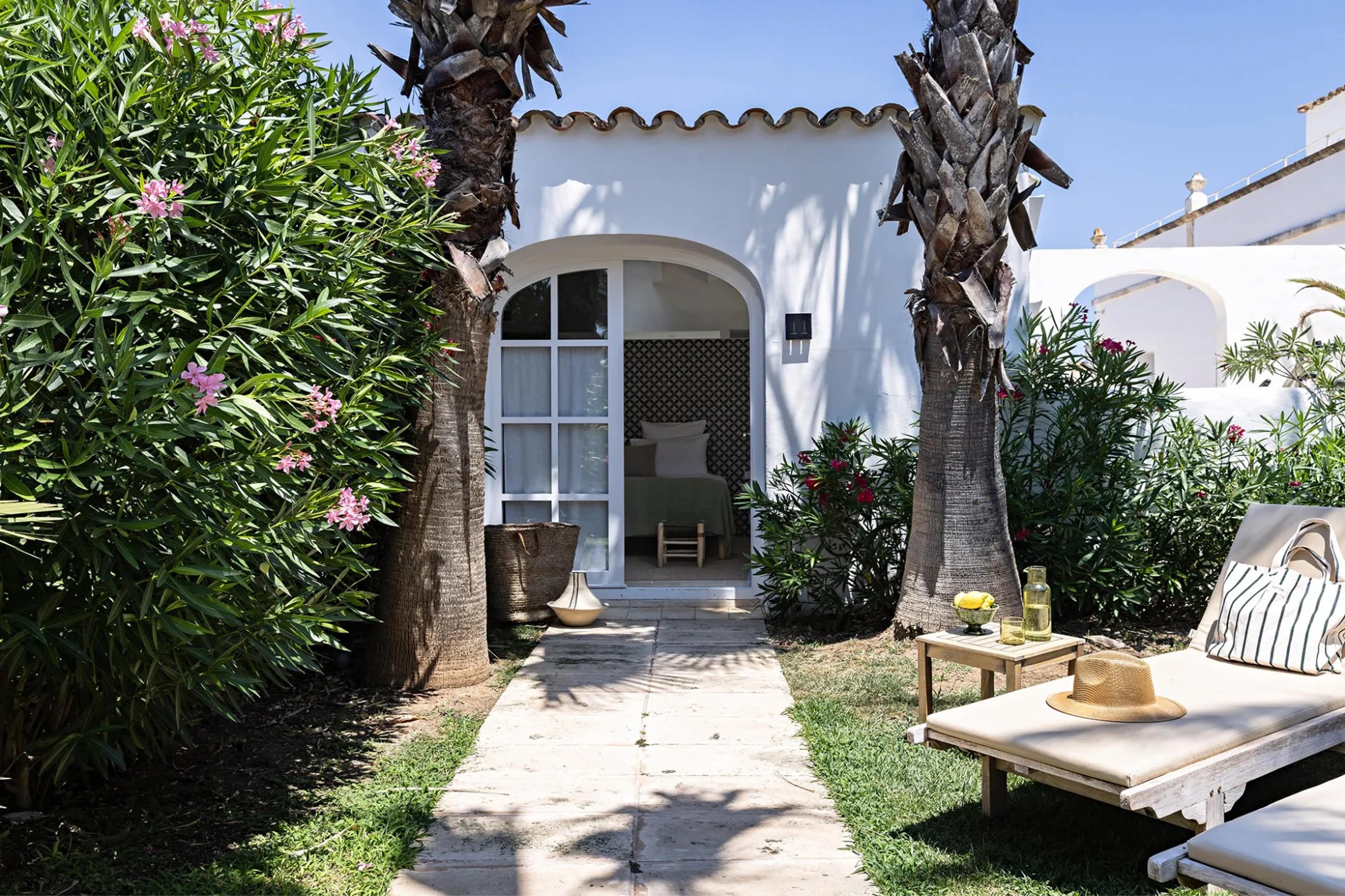a stone path through a garden of pink oleander and palm trees leading to the open arched door of a whitewashed building, with a sun lounger and a straw hat in the foreground