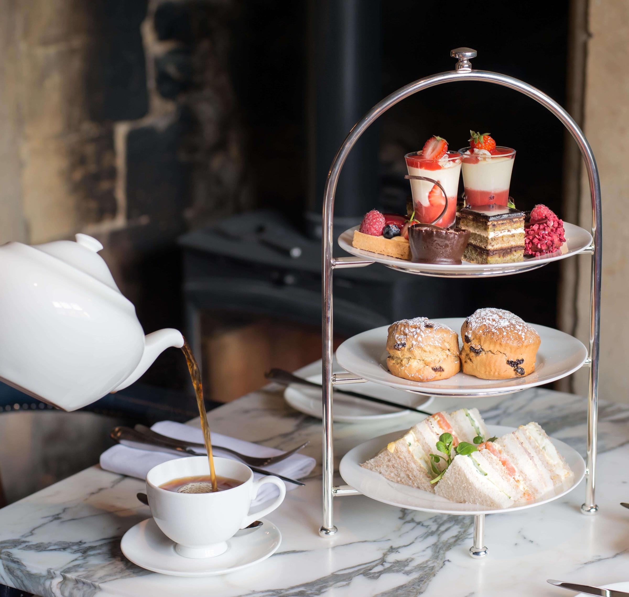 A three-tiered stand of finger sandwiches, scones and small desserts on a marble table, with tea being poured into a cup from a white teapot.