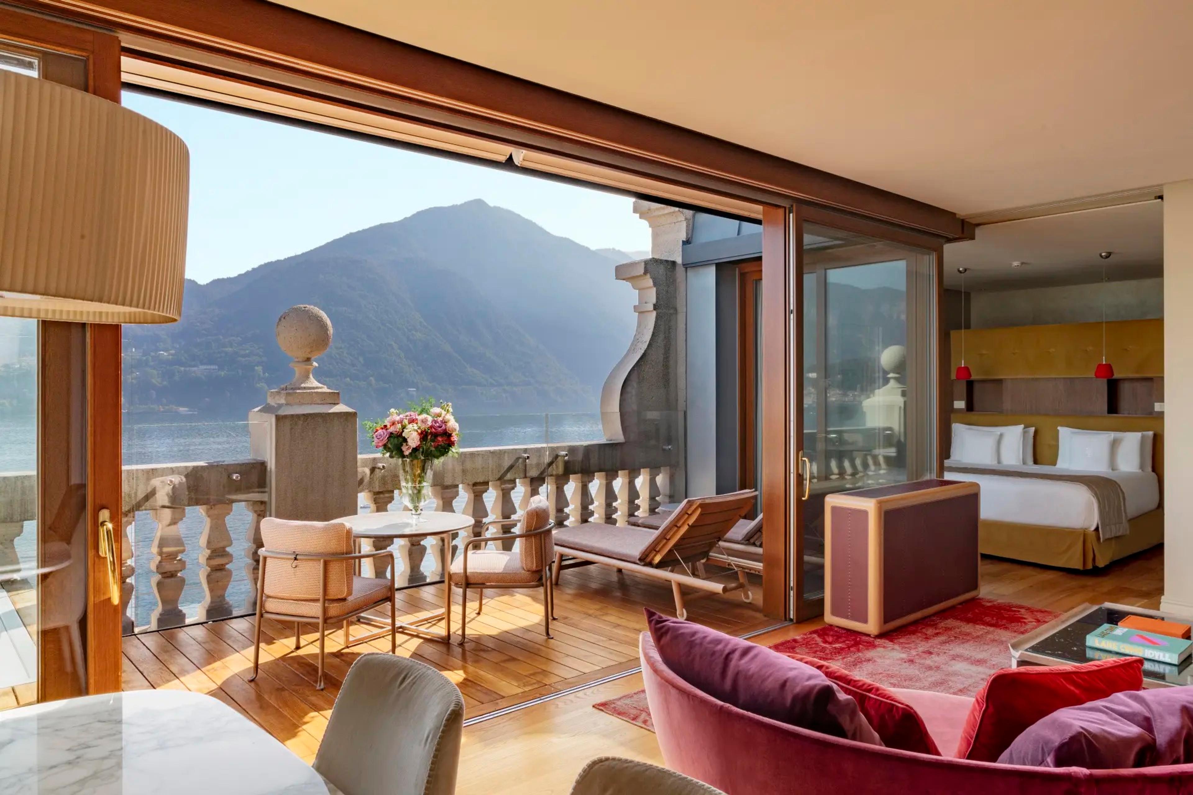 A hotel suite opens through glass doors to a balcony with a dining table and lounger overlooking the lake and mountains, a bed and pink sofa visible inside.