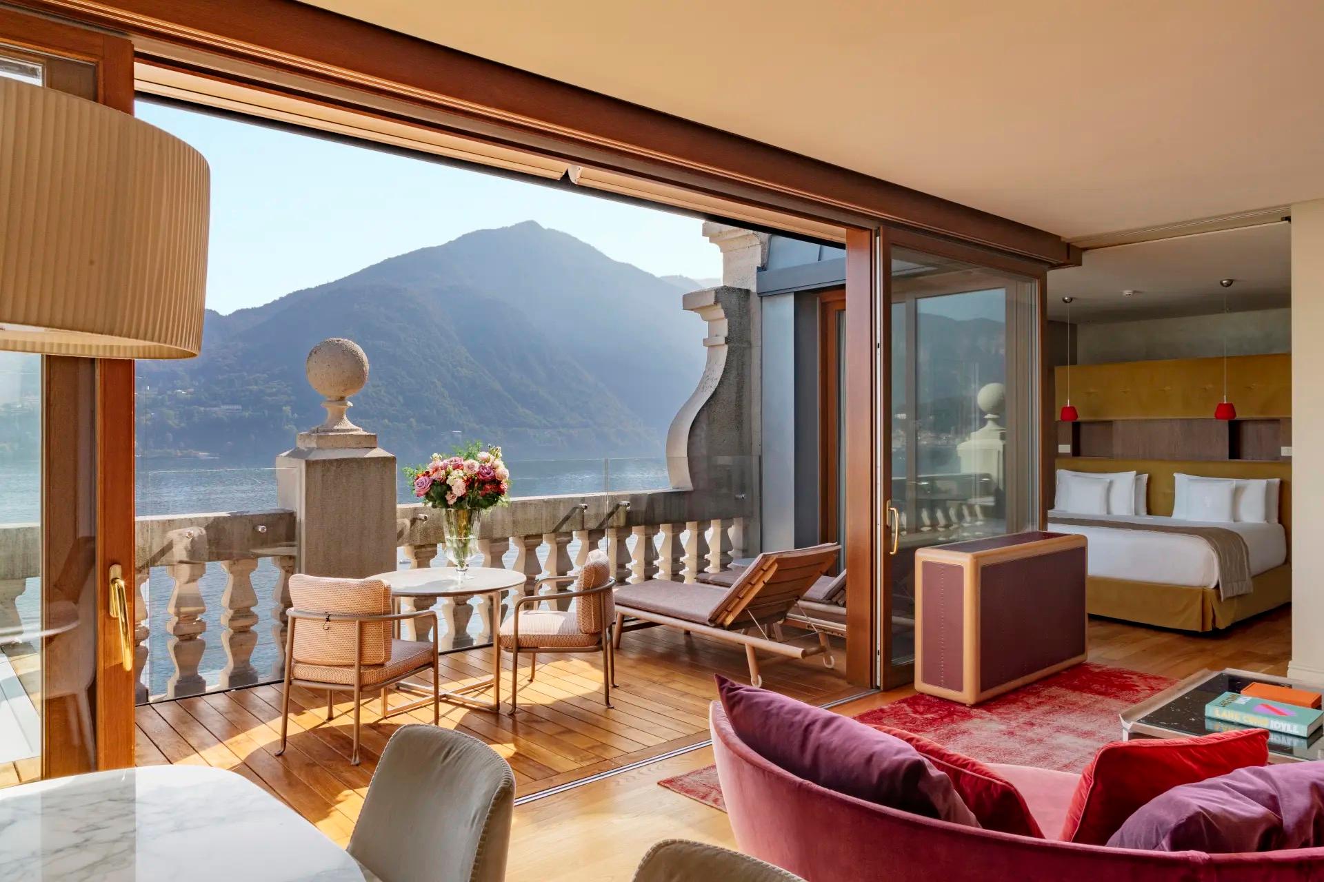 A hotel suite opens through glass doors to a balcony with a dining table and lounger overlooking the lake and mountains, a bed and pink sofa visible inside.