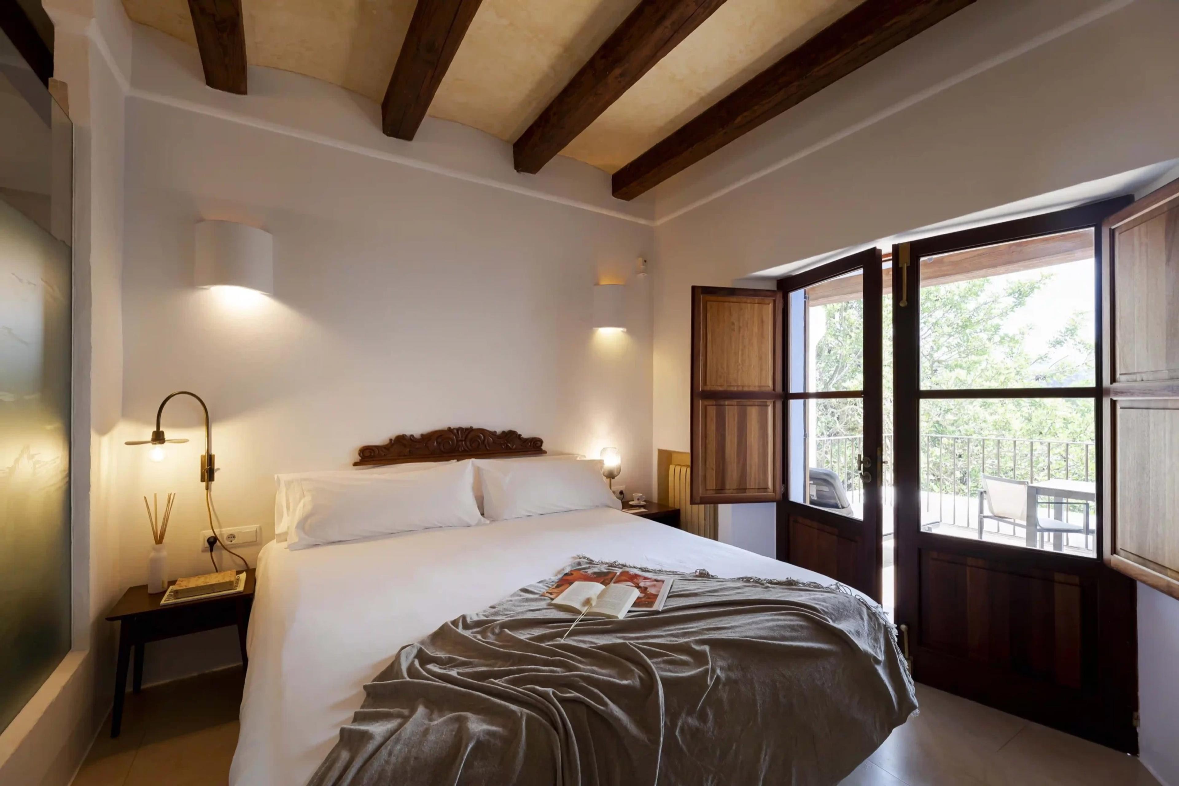 A bedroom with a carved wooden headboard, exposed ceiling beams, and open shutters onto a small balcony with a table and chairs.