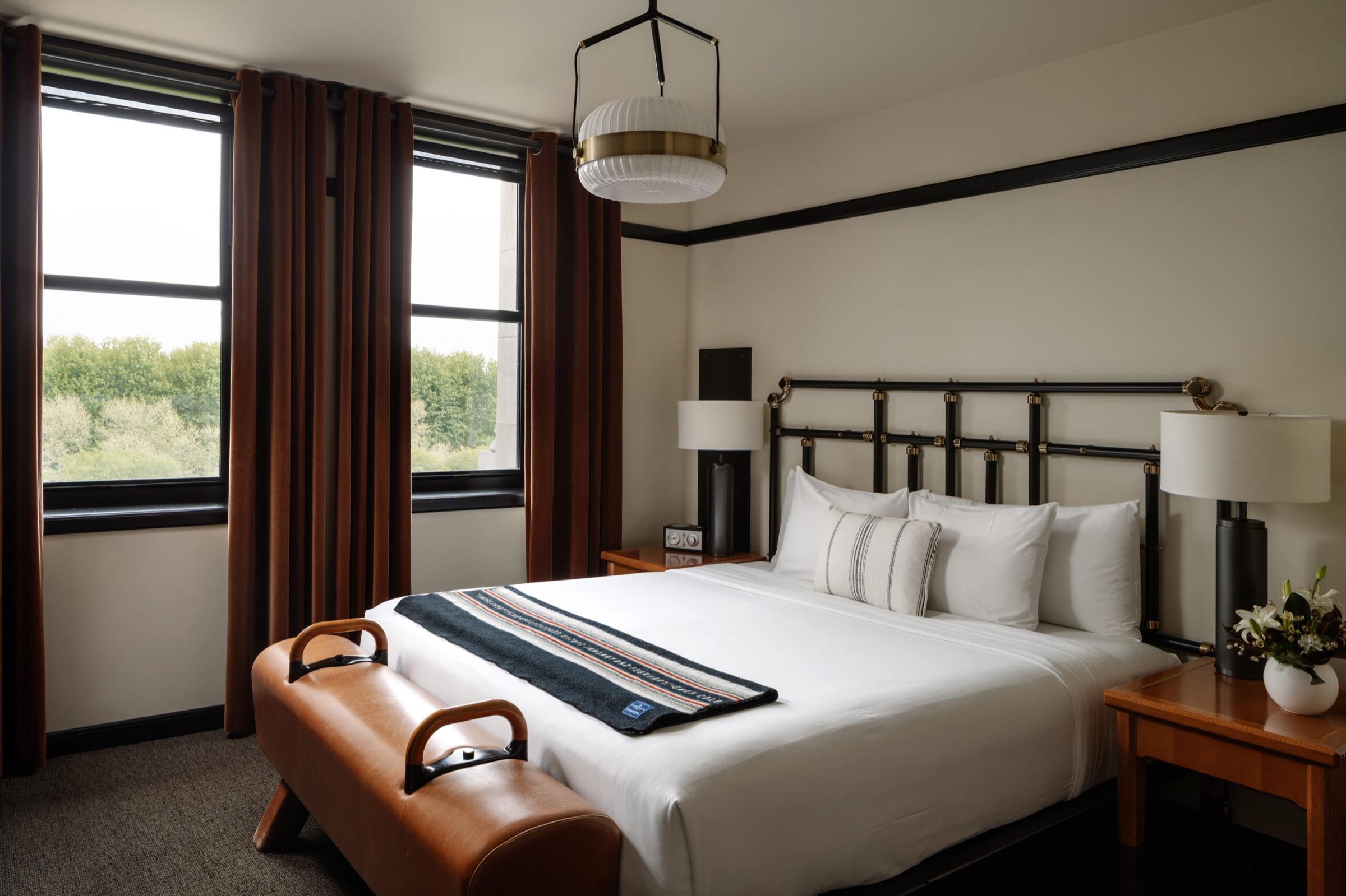 A king bed with a leather bench at its foot, flanked by nightstands with lamps and a vase of flowers, with rust colored curtains and trees visible through the windows.