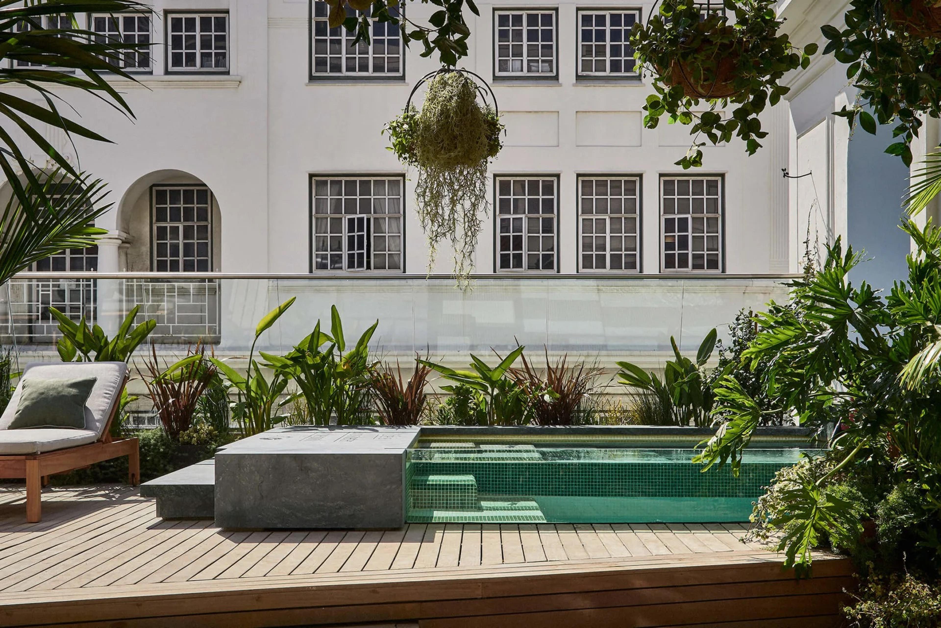 A small green-tiled plunge pool set into a timber deck, with a stone step beside it, planting around the edge and a white building with many windows behind.