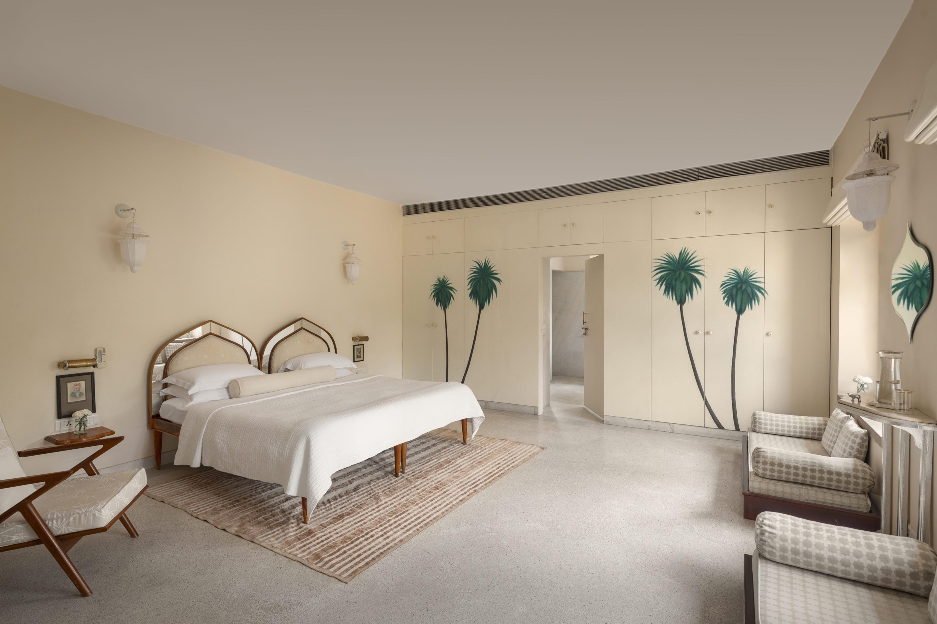 A bedroom with a mirrored headboard, a reading chair and a wardrobe painted with a palm tree, white bed linen and a striped rug.