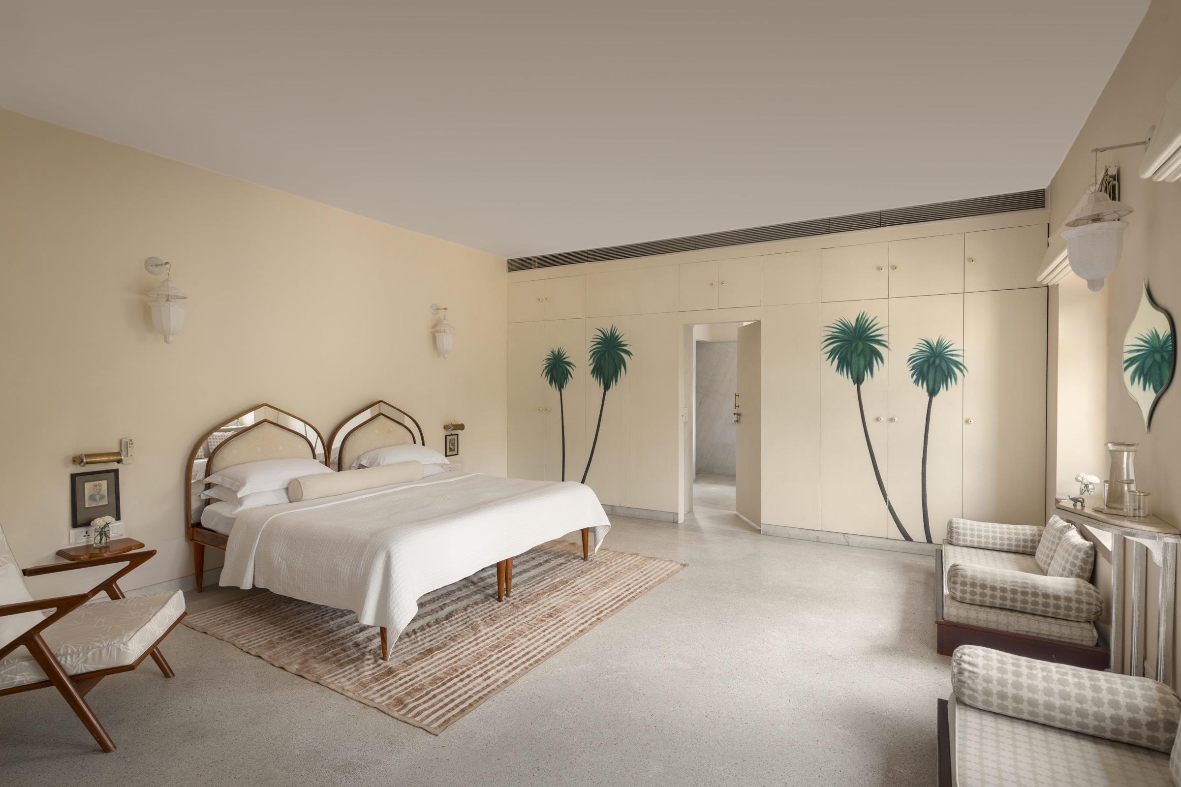 A bedroom with a mirrored headboard, a reading chair and a wardrobe painted with a palm tree, white bed linen and a striped rug.