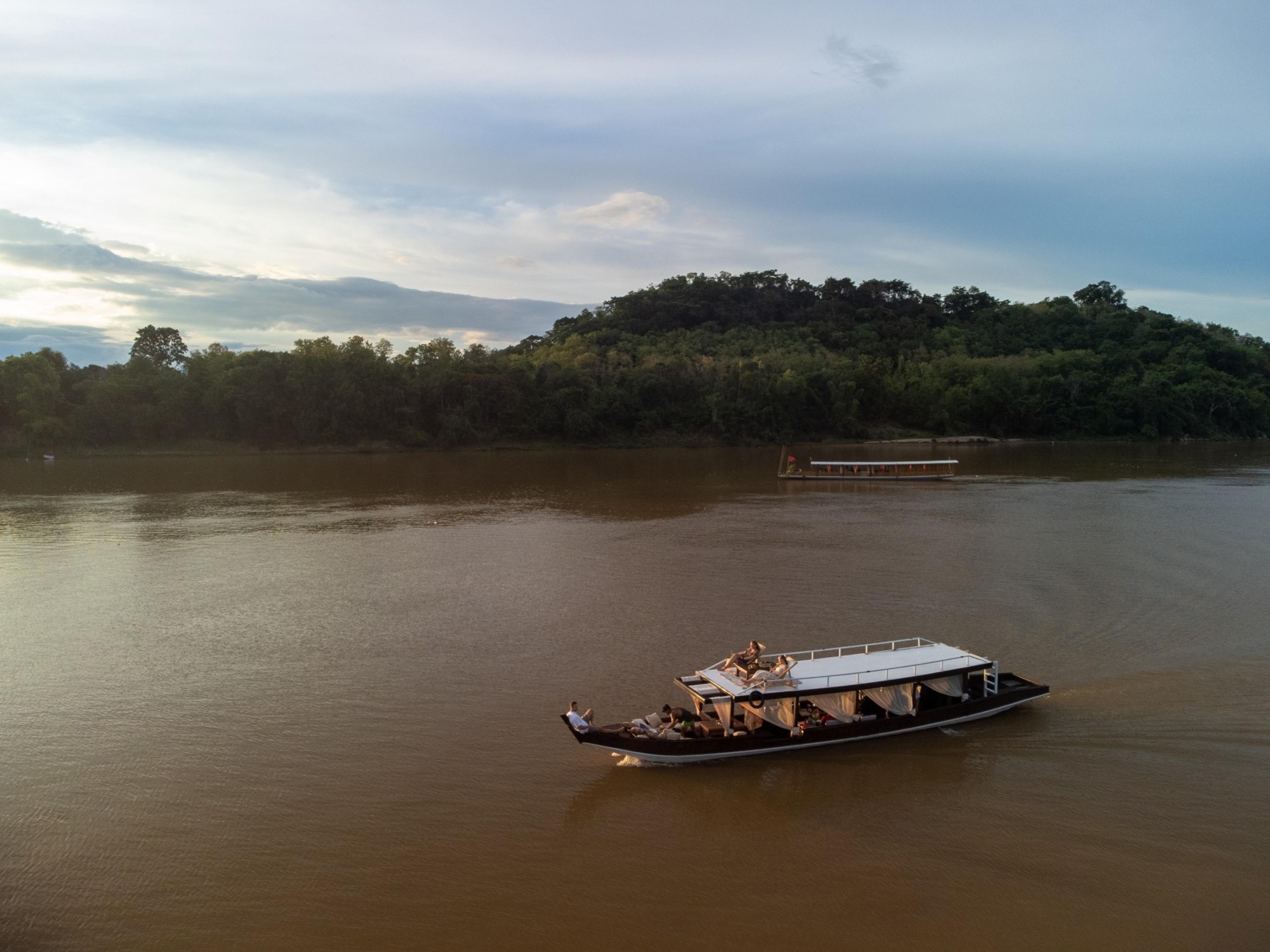 An aerial view at dusk of two traditional wooden boats on a wide brown river, with forested hills rising behind.