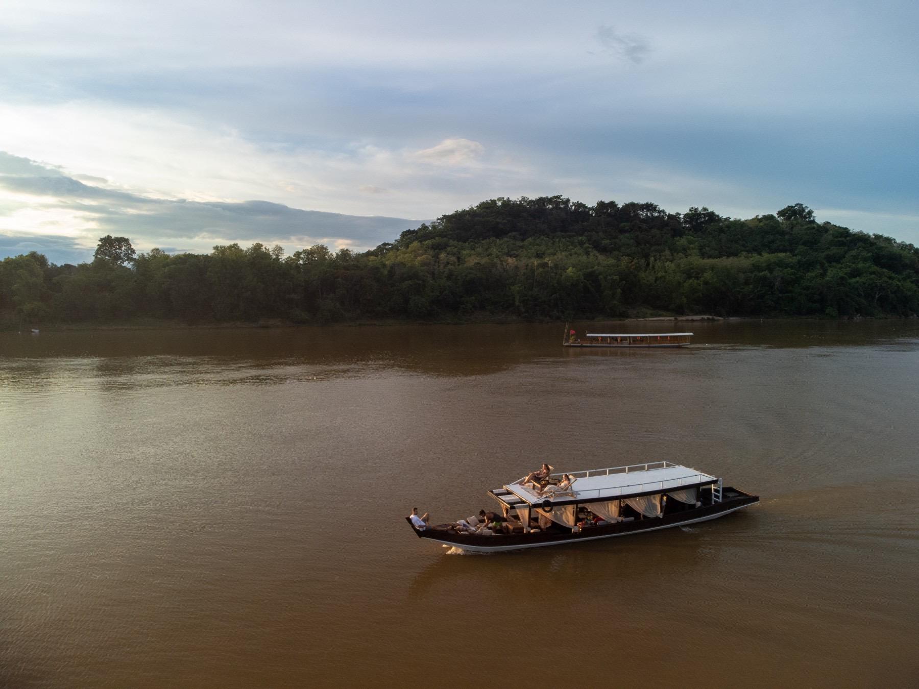 An aerial view at dusk of two traditional wooden boats on a wide brown river, with forested hills rising behind.