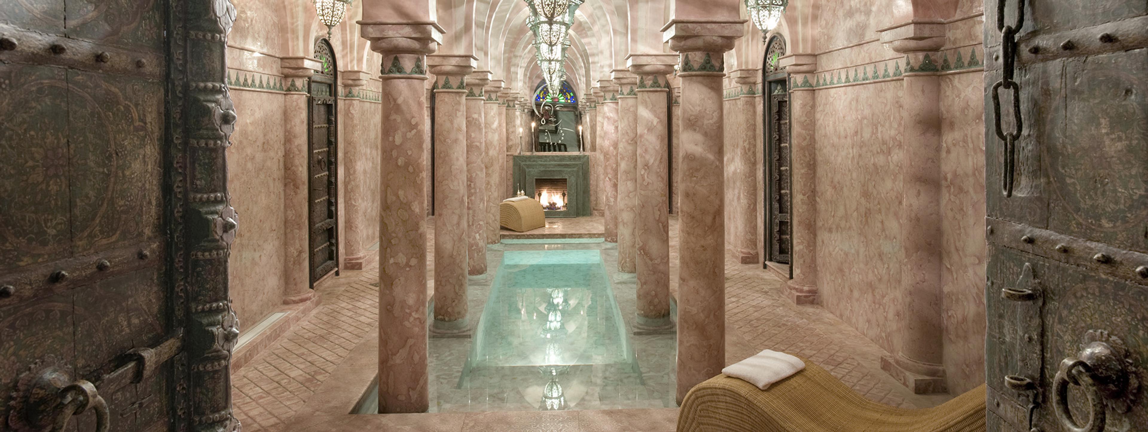 A columned corridor with a row of sunken turquoise plunge pools down the centre, lit by hanging lanterns, ending at a fireplace.