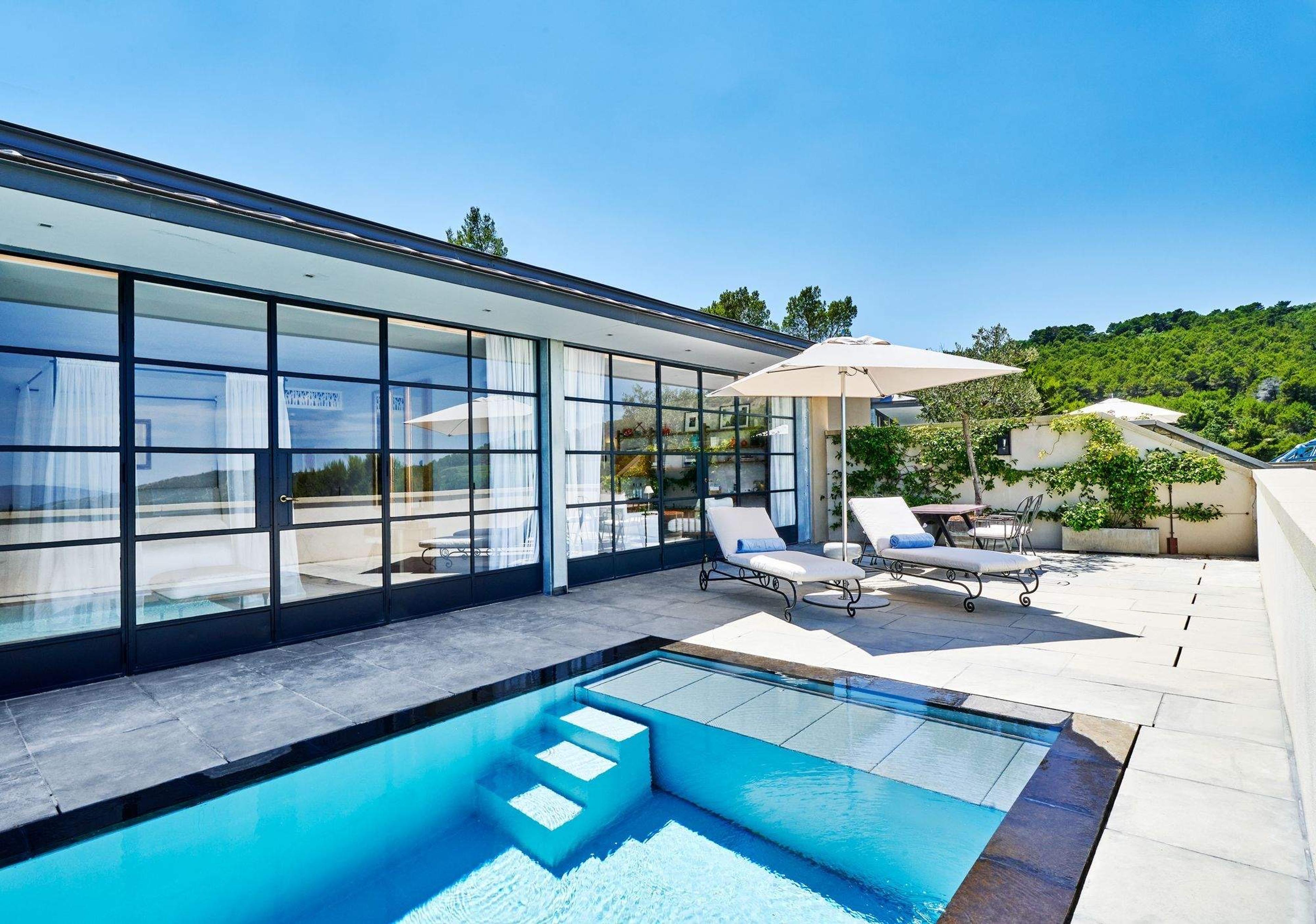 A narrow pool with stone steps leading into the water beside a glass-fronted villa, with two sun loungers and a cream umbrella on the terrace.