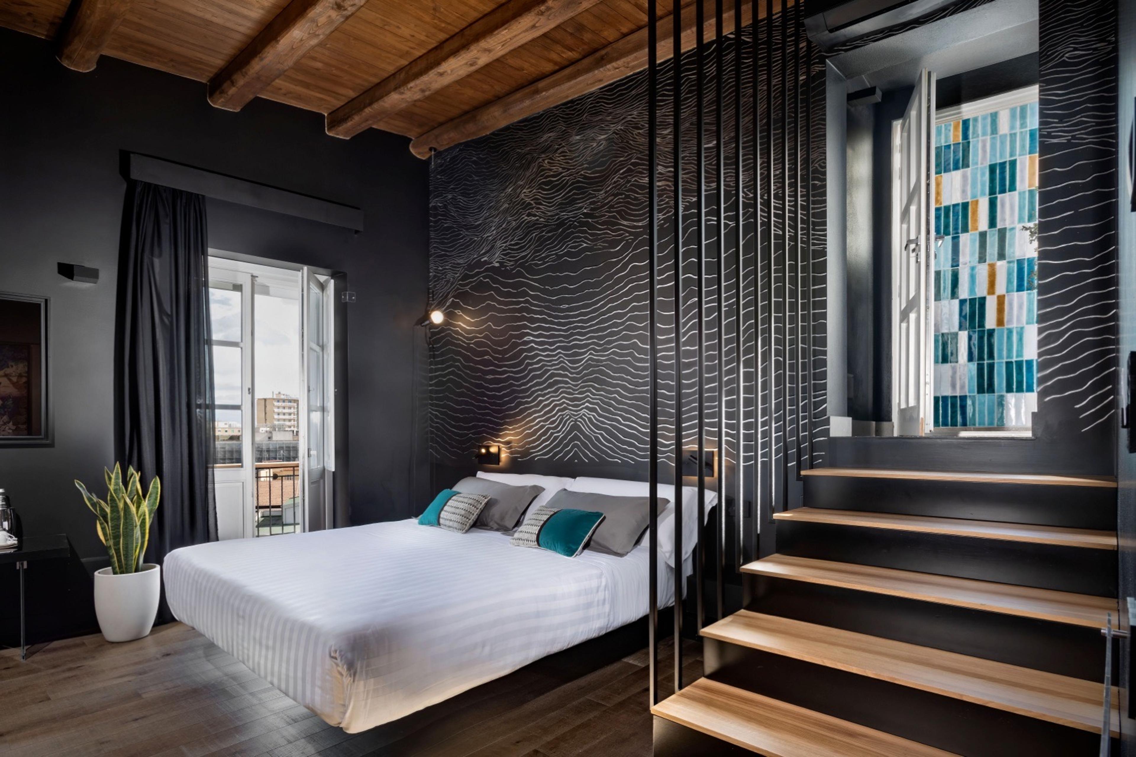 A dark bedroom with a patterned wall behind the bed, an open-tread staircase leading up past a stained glass window, and a potted plant by the curtained doors.
