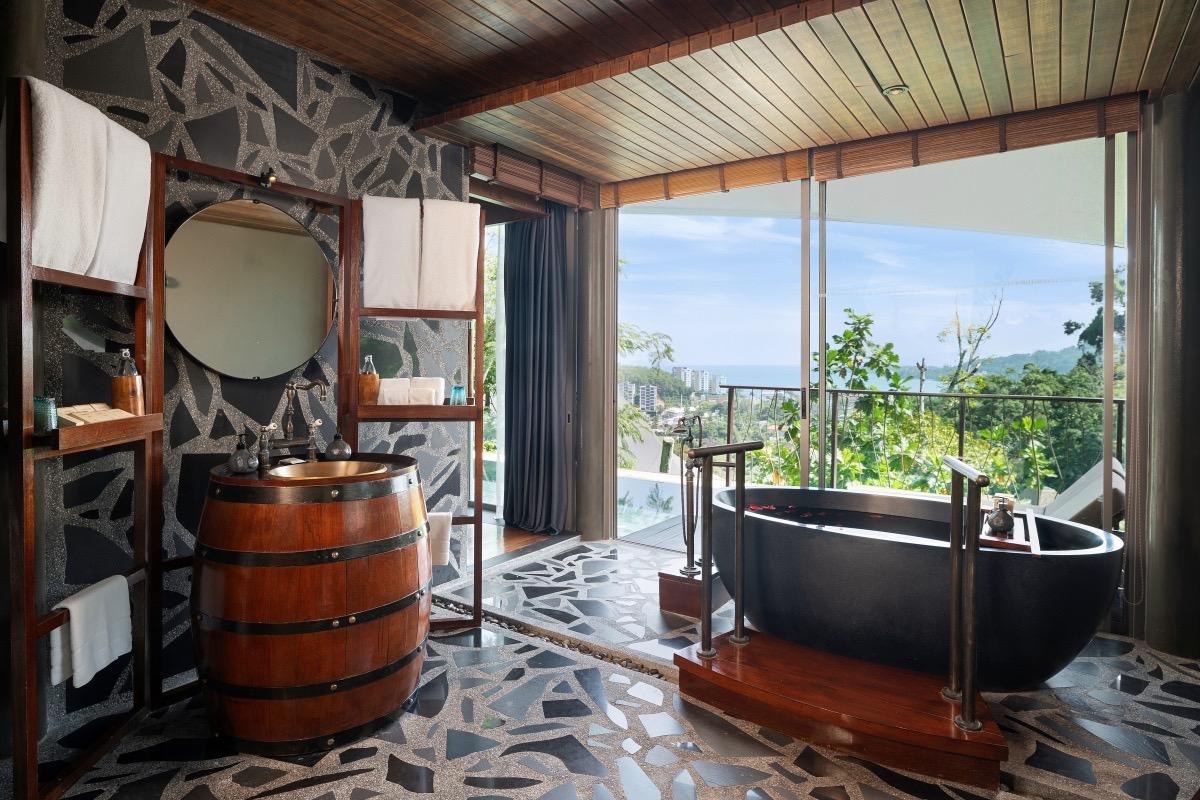 A round black bathtub on a wooden platform beside floor-to-ceiling windows showing a coastal view, with a barrel-shaped sink nearby.