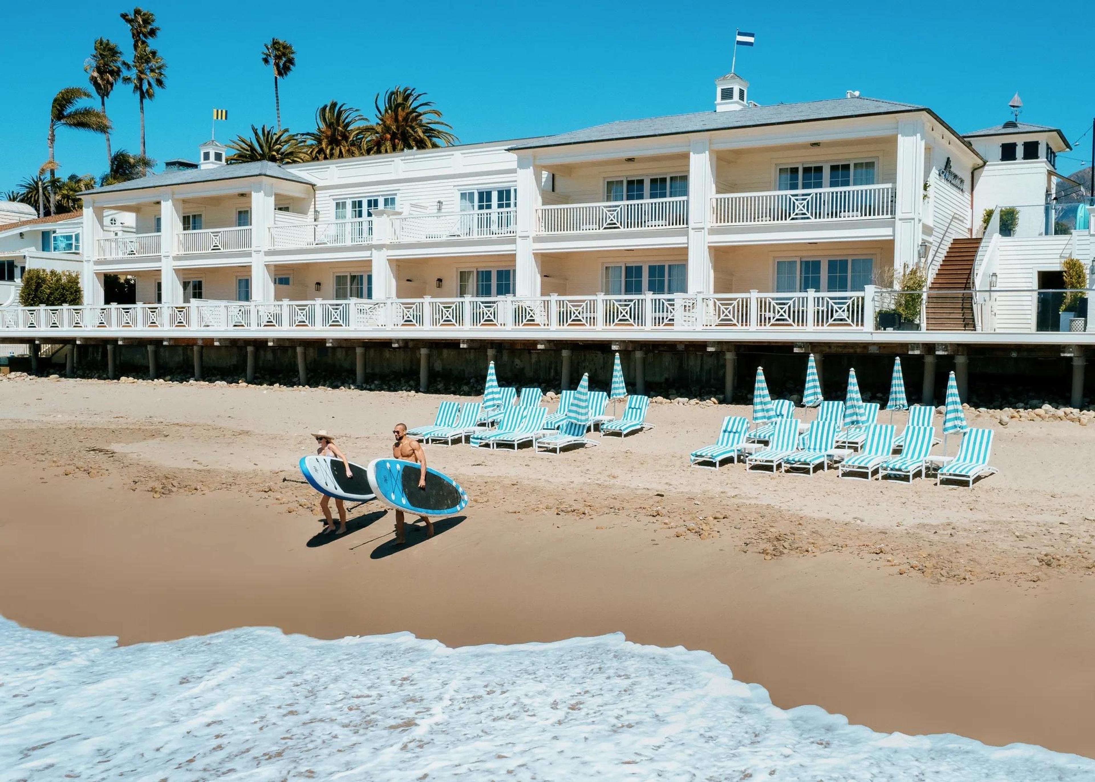 Two people carrying surfboards along a beach in front of a white clapboard hotel, with striped loungers and umbrellas set out on the sand.