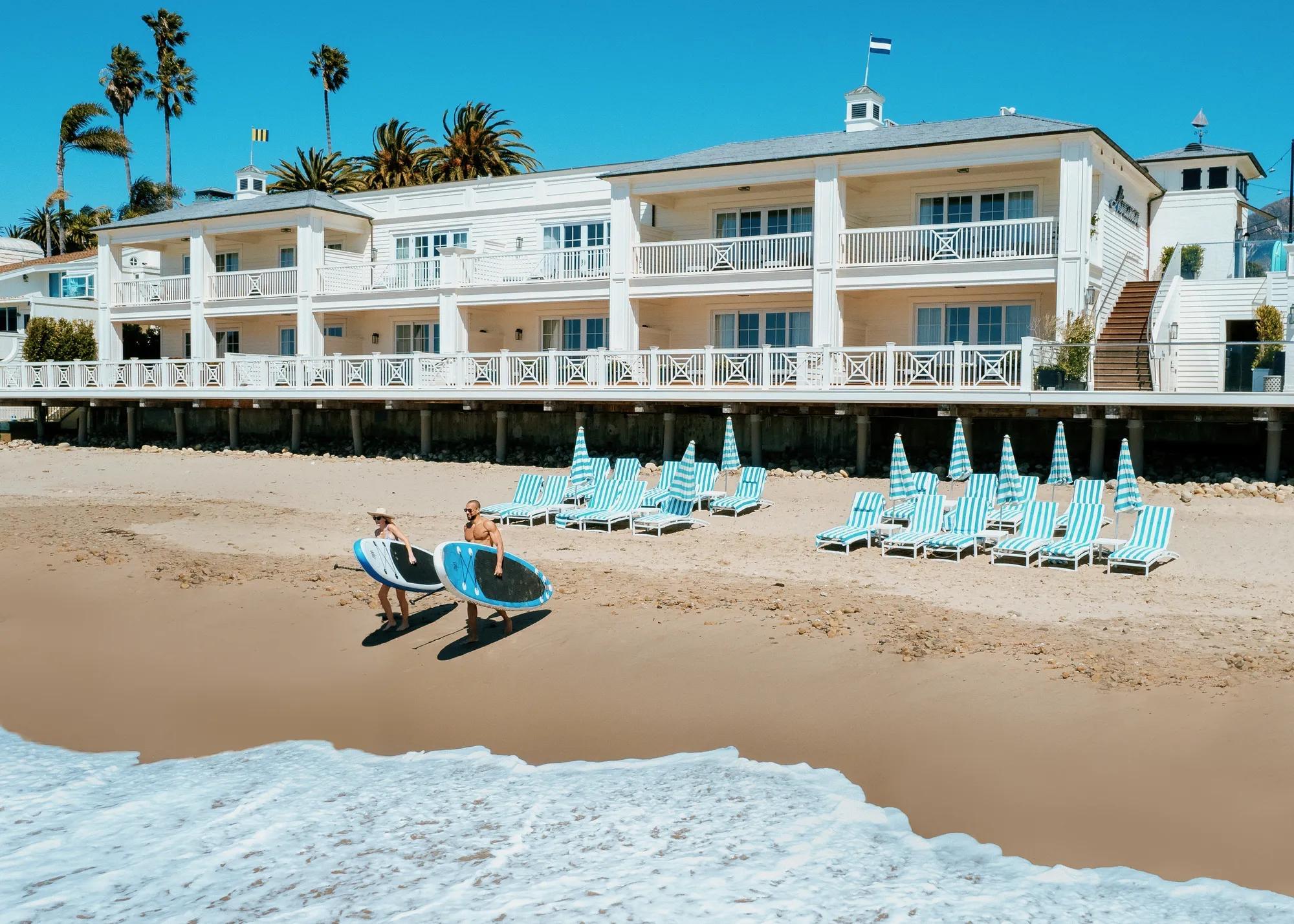 Two people carrying surfboards along a beach in front of a white clapboard hotel, with striped loungers and umbrellas set out on the sand.