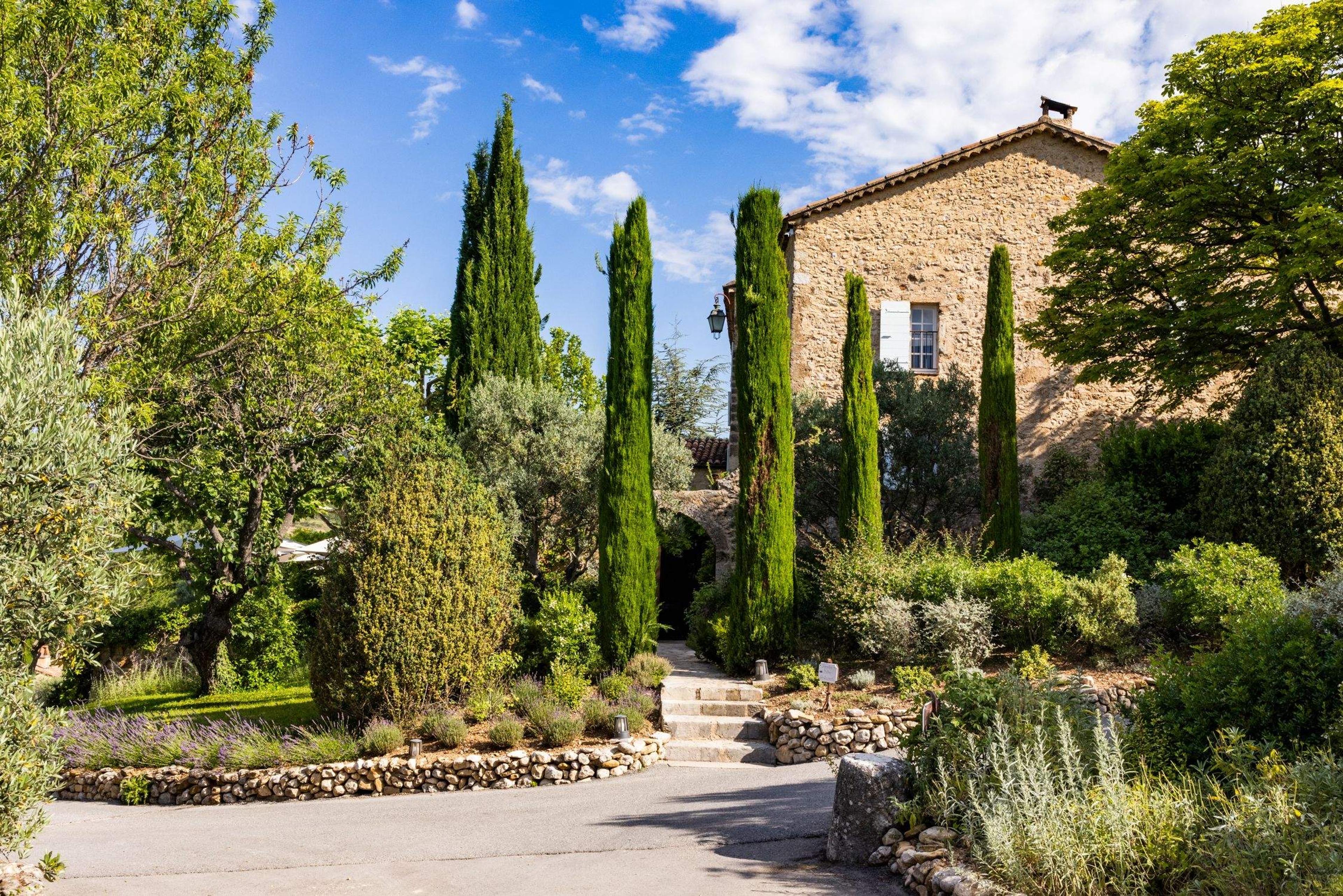 A gravel driveway lined with tall cypress trees leading up to a two-storey stone farmhouse under a blue sky with scattered clouds.
