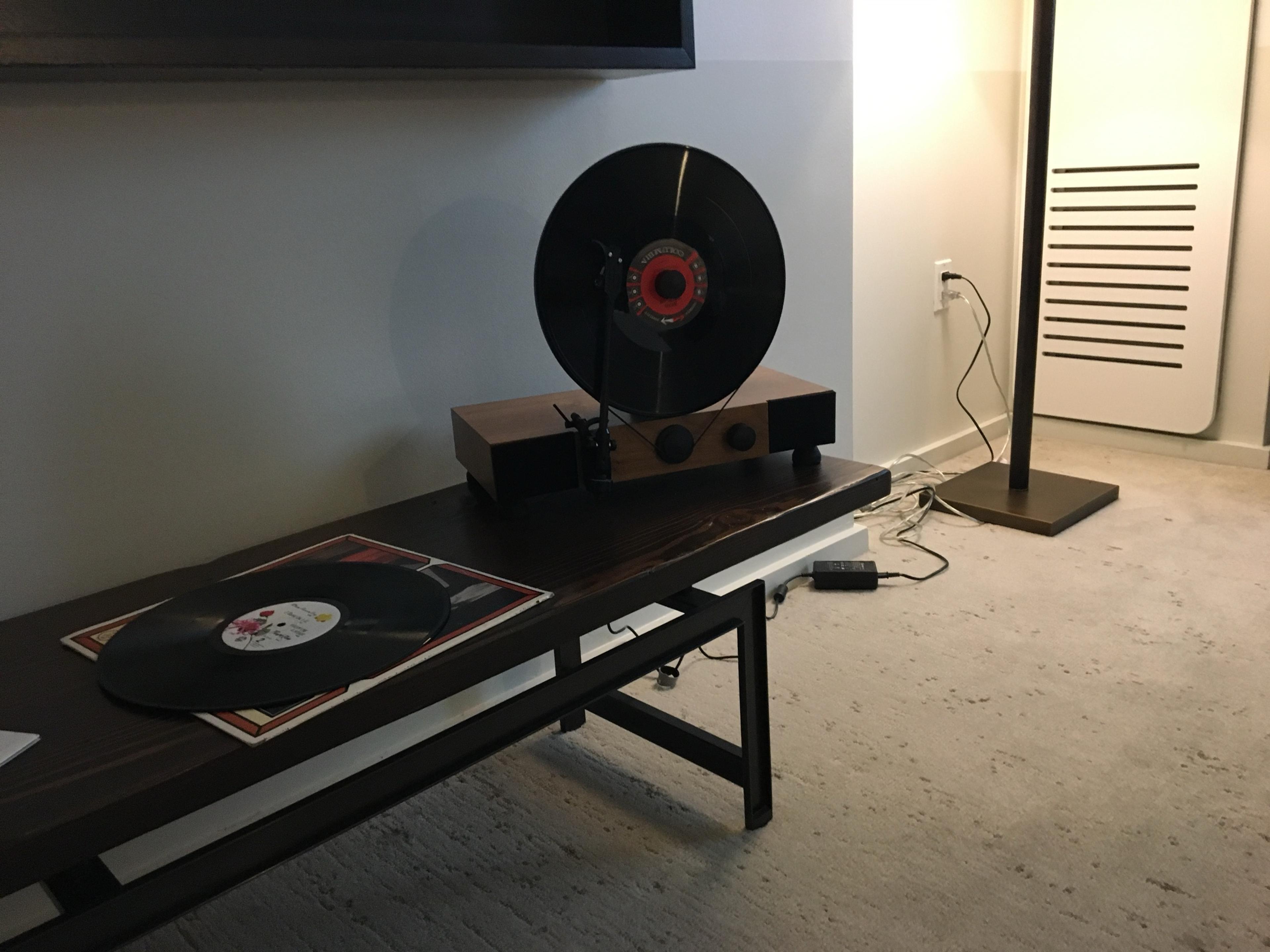 A record player standing upright on a low wooden table in a hotel room, with a record leaning beside it