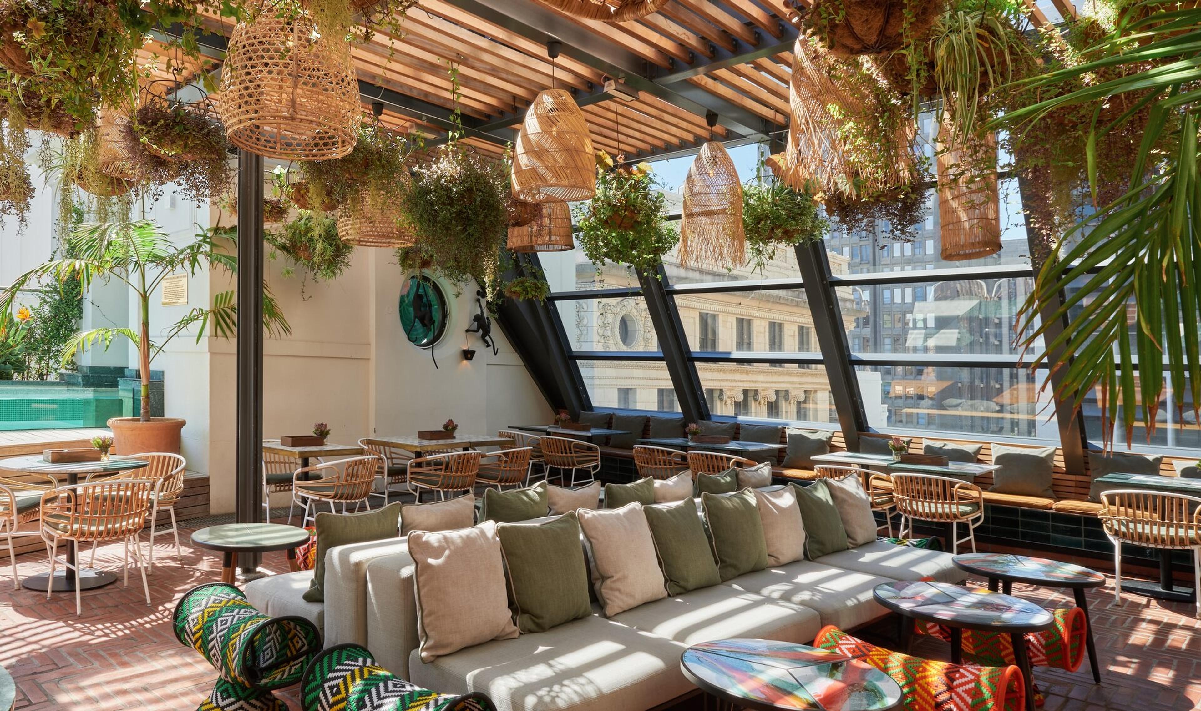 A covered rooftop terrace with a timber pergola, wicker pendant lamps and hanging plants above low sofas and tables, with glass walls looking out over the facades of city buildings.