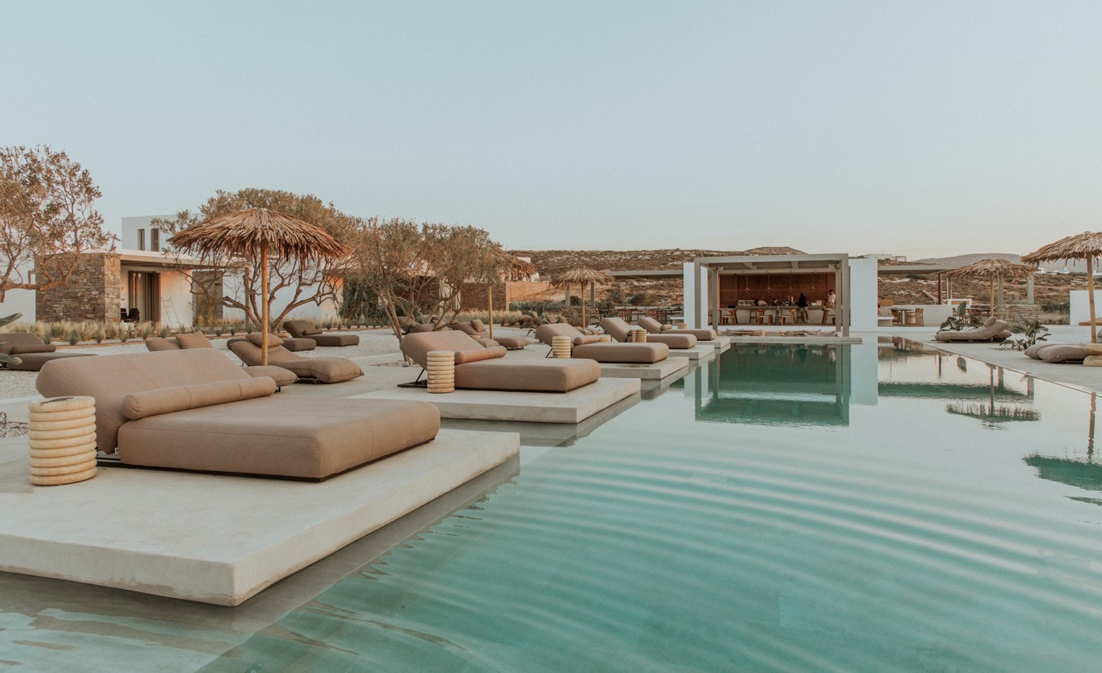 Rows of sun loungers beside a long pool at dusk, leading to a lit pavilion with hills behind.