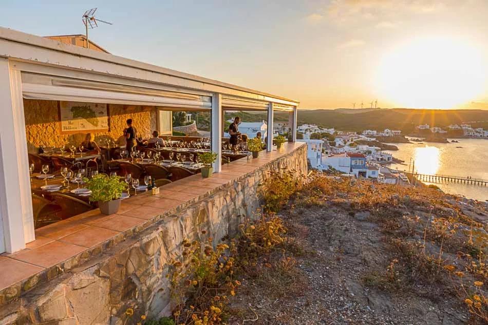 A covered terrace of tables on rocks above the sea, at sunset.
