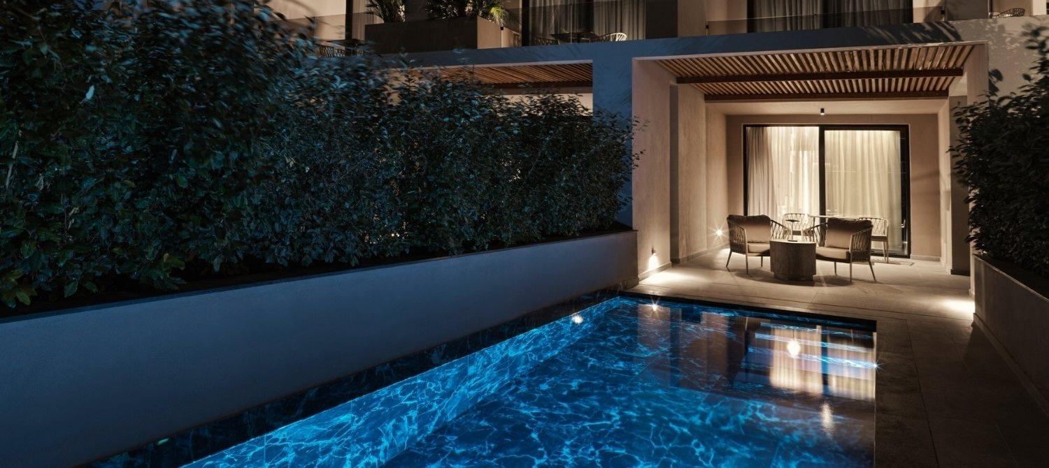 A private plunge pool lit blue at night beside a terrace with armchairs and a lit suite behind glass doors.