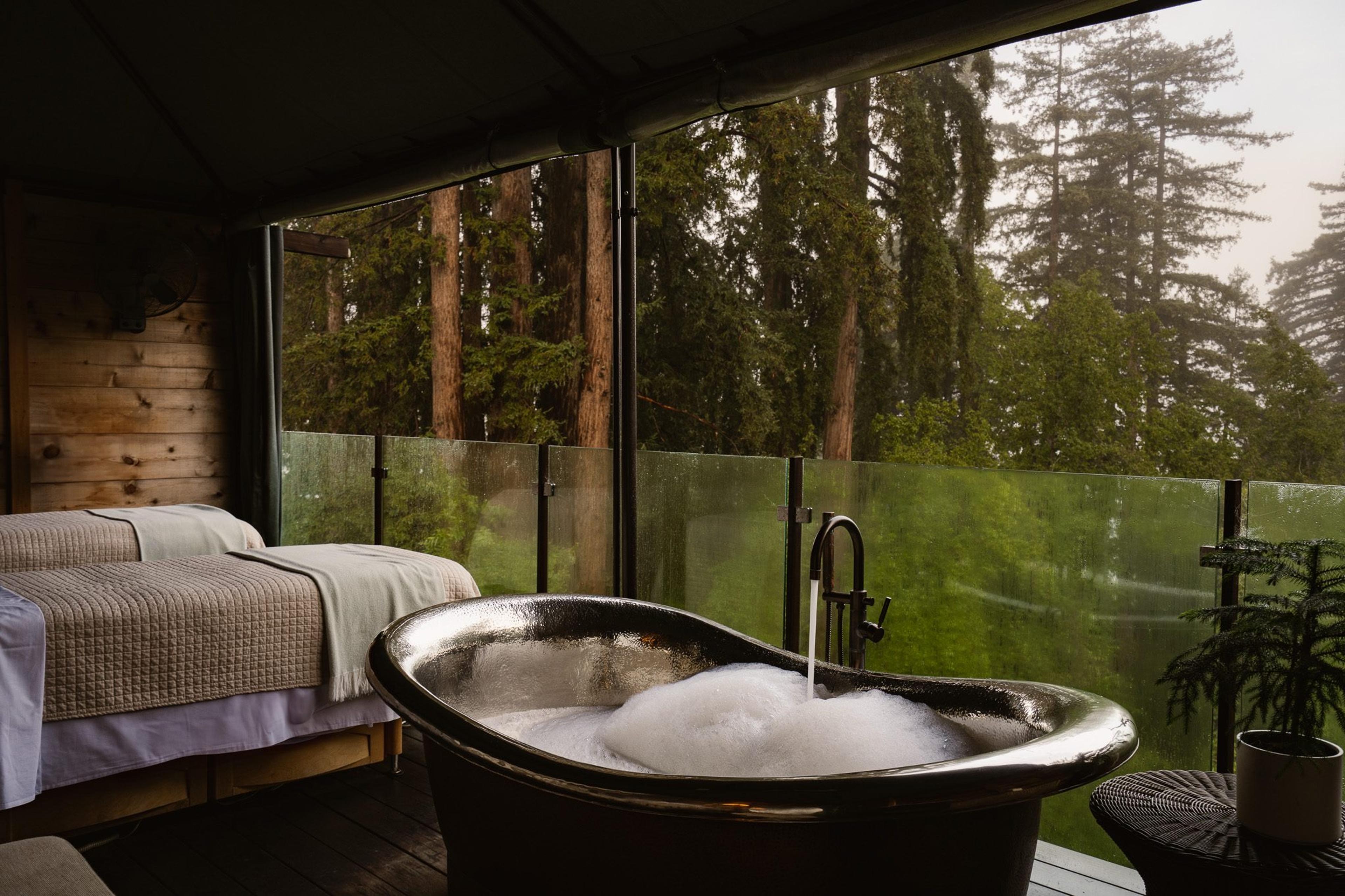 a freestanding bathtub full of bubbles on a deck, with twin beds and redwood trees visible through a glass screen