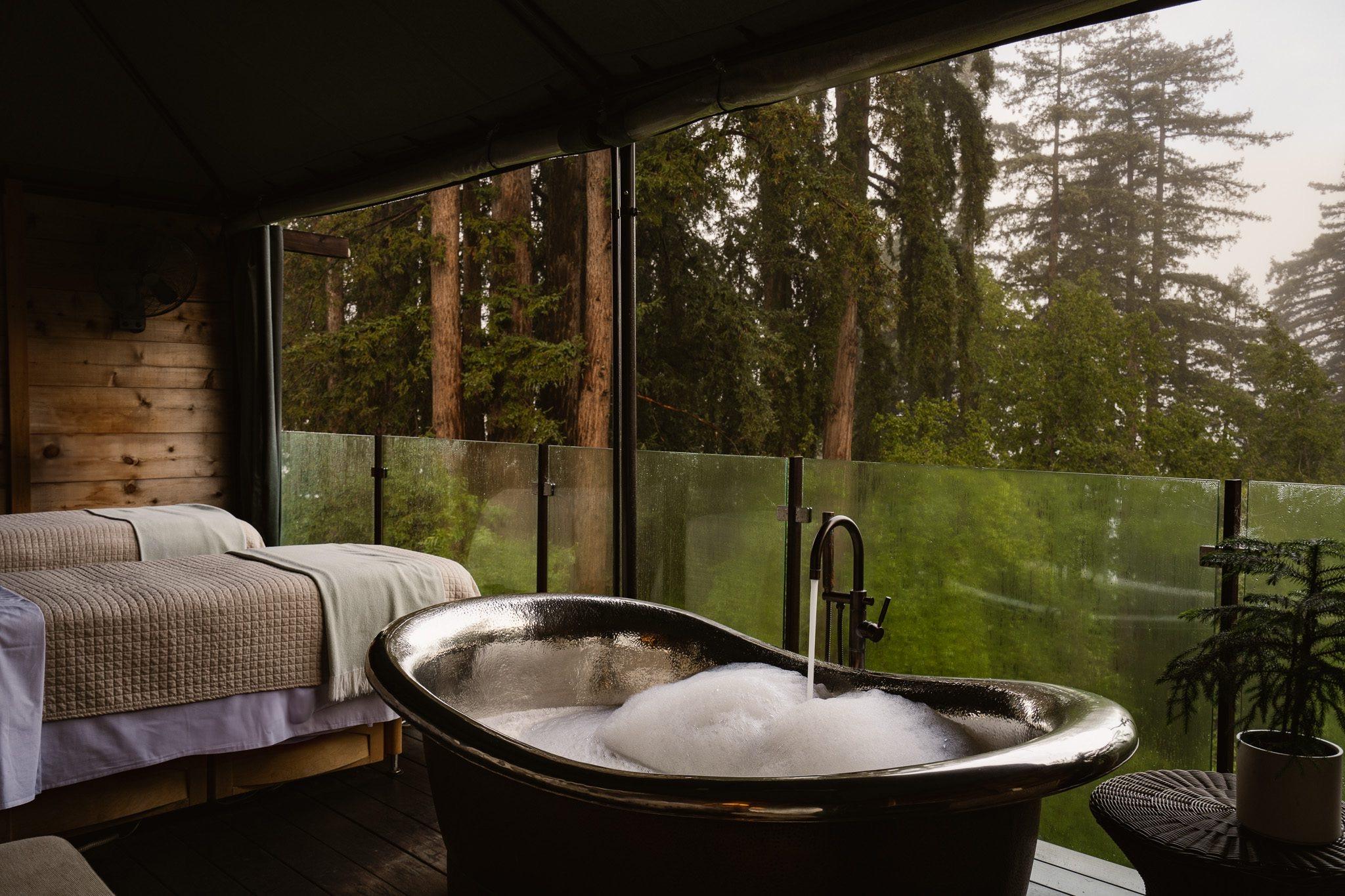 a freestanding bathtub full of bubbles on a deck, with twin beds and redwood trees visible through a glass screen