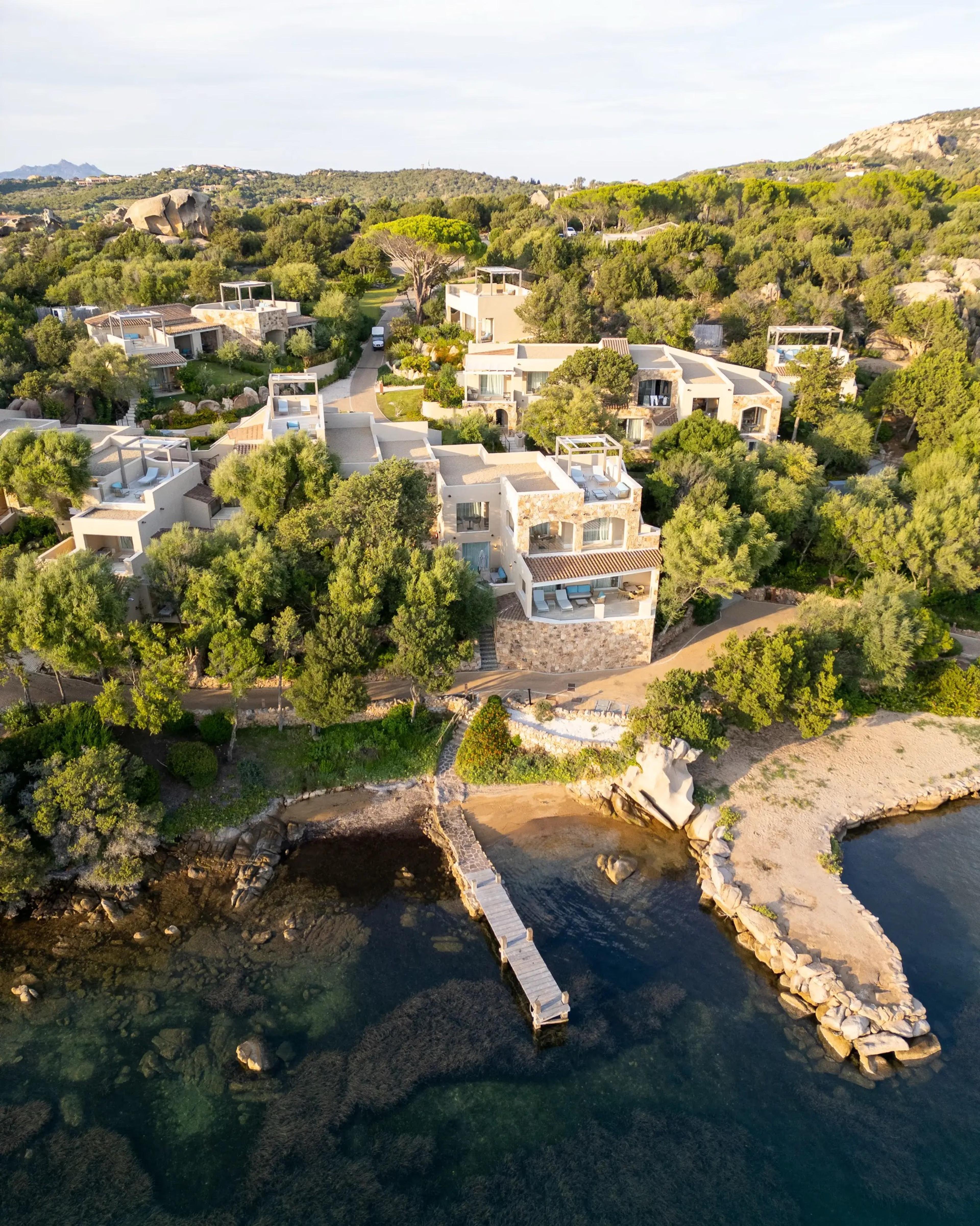 An aerial view of a cluster of stone and white villas among pine trees above a rocky cove with a wooden pier reaching into clear water.