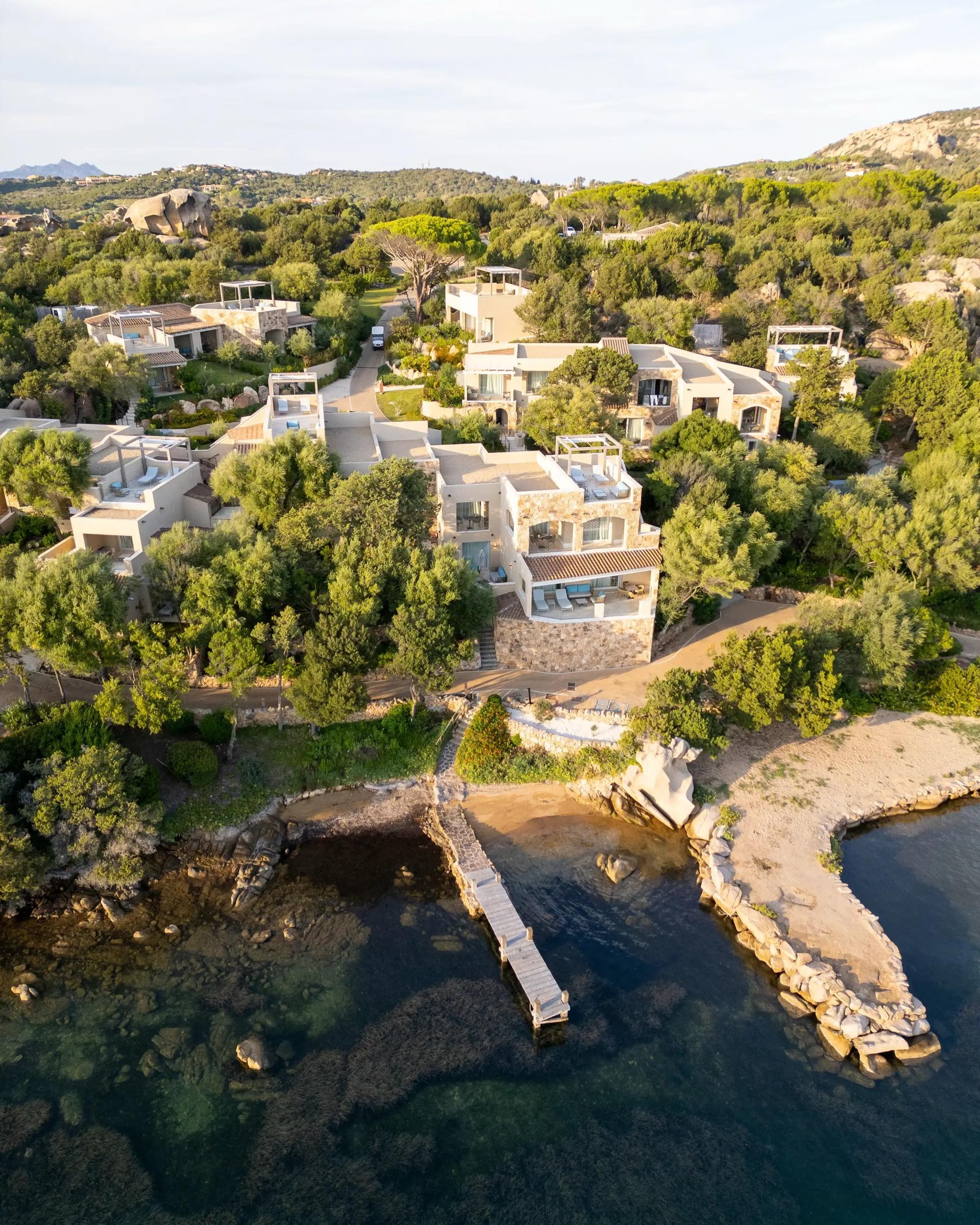 An aerial view of a cluster of stone and white villas among pine trees above a rocky cove with a wooden pier reaching into clear water.
