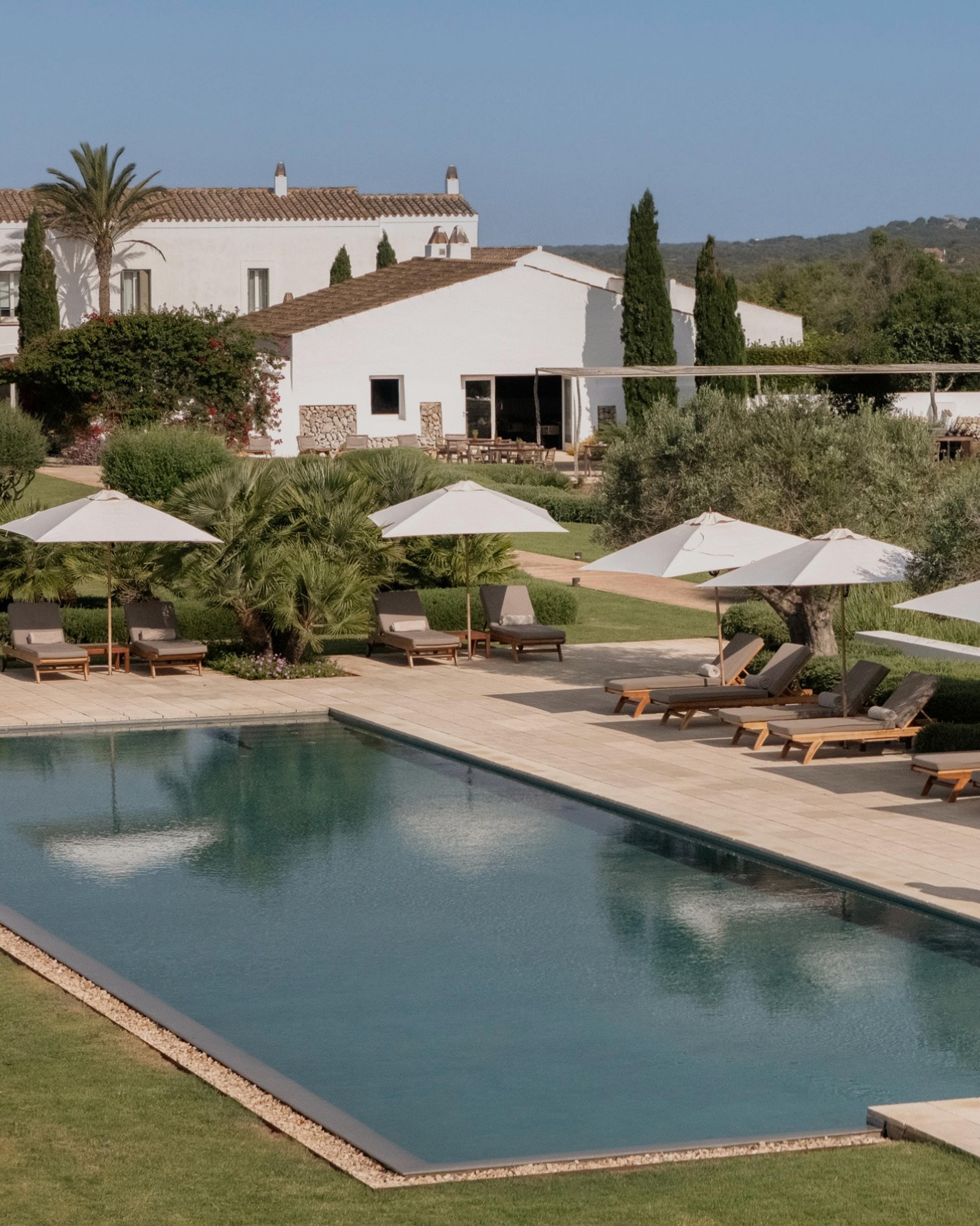A swimming pool with sun loungers and white umbrellas beside a white building with a tiled roof, seen from above with trees and hills beyond.