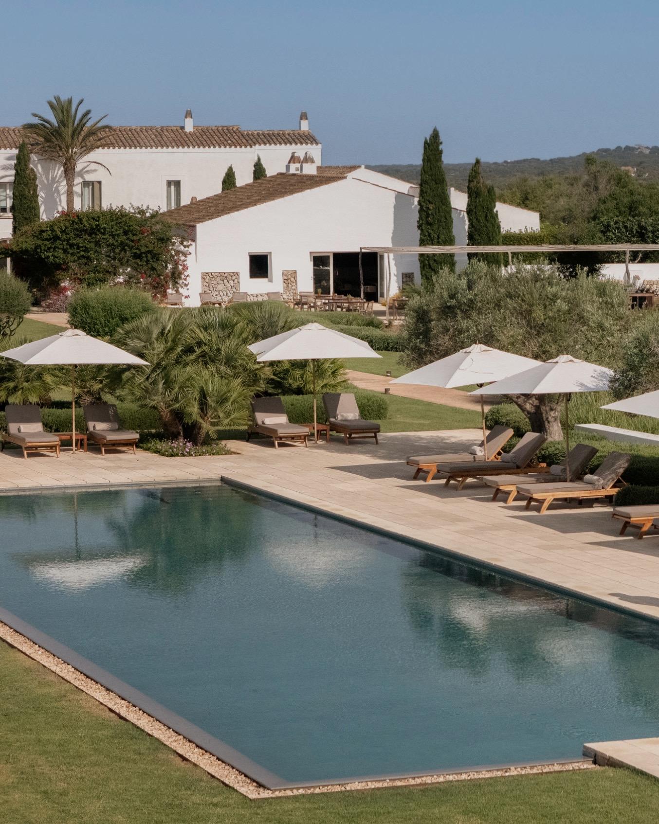 A swimming pool with sun loungers and white umbrellas beside a white building with a tiled roof, seen from above with trees and hills beyond.