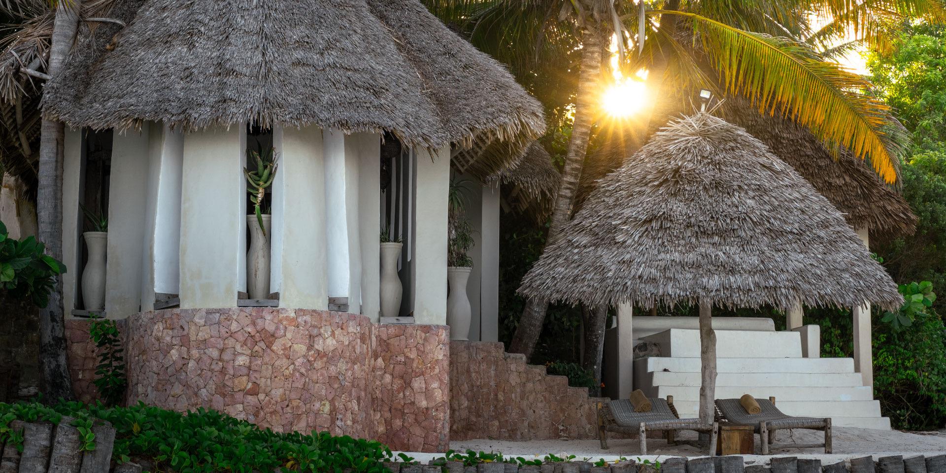 Thatched-roof bungalows with whitewashed walls and a stone base, lit by low sun through the palms.