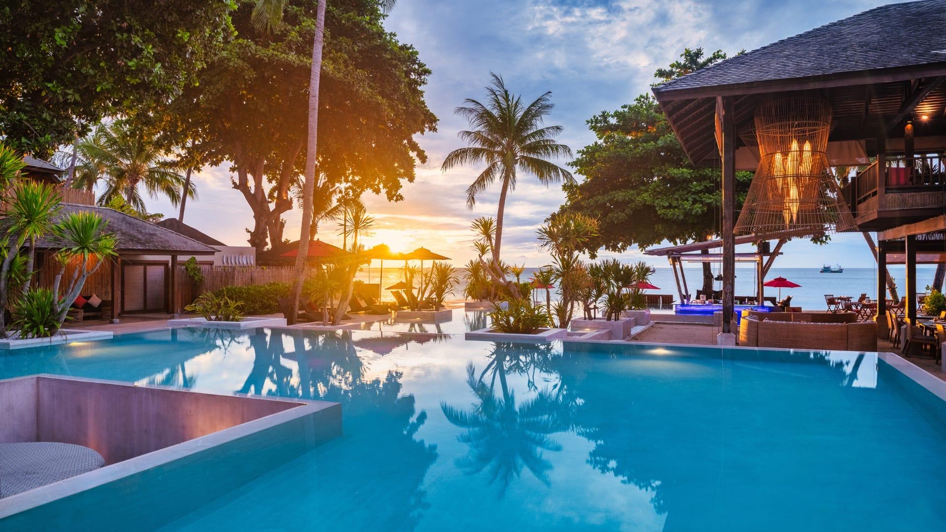 An infinity pool reflecting a sunset through palm trees, with a wooden restaurant building and the sea beyond.