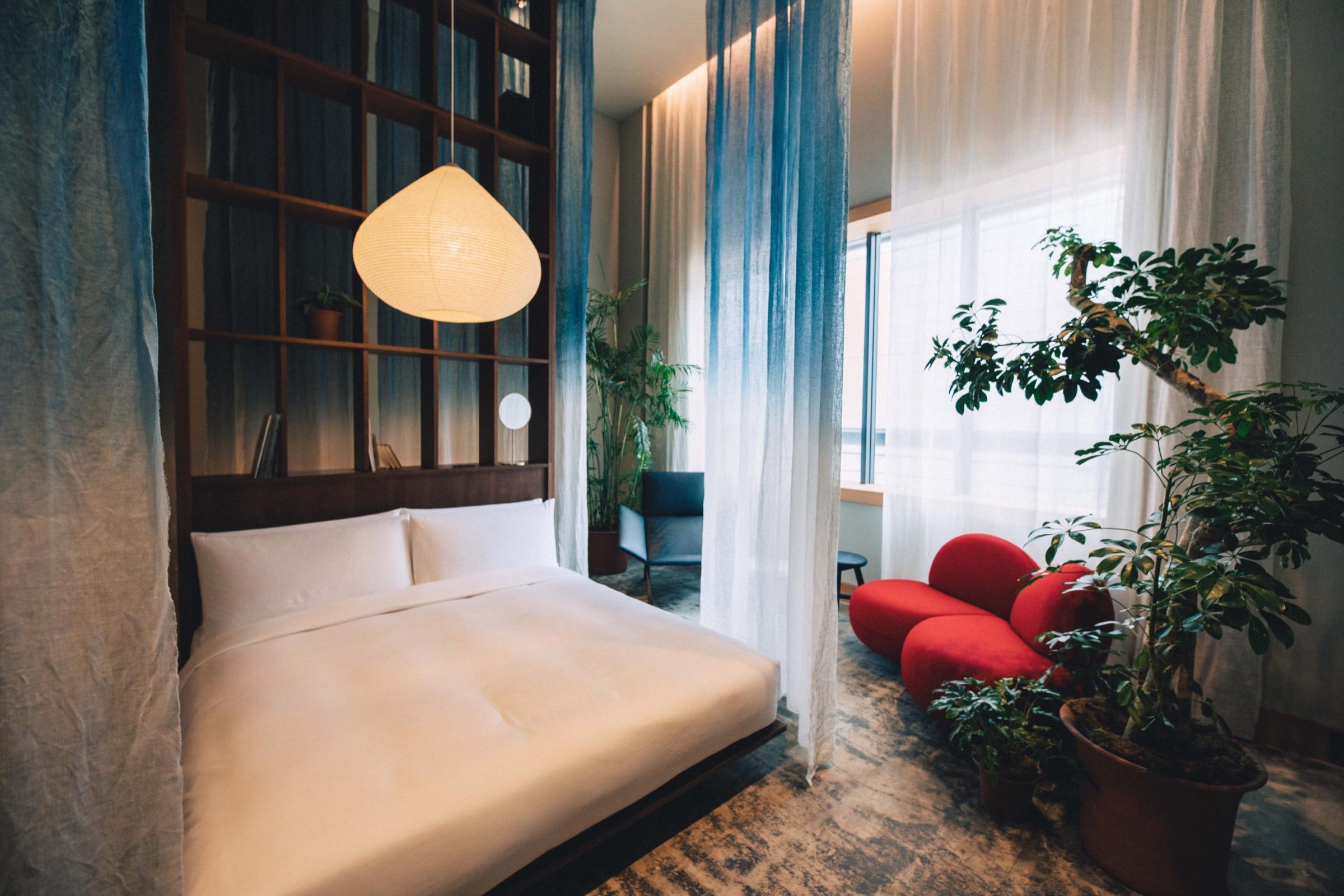 A bedroom with a dark wood headboard and shelf, a white bed under a paper lantern, blue dip-dyed curtains, and a red curved sofa by the window.