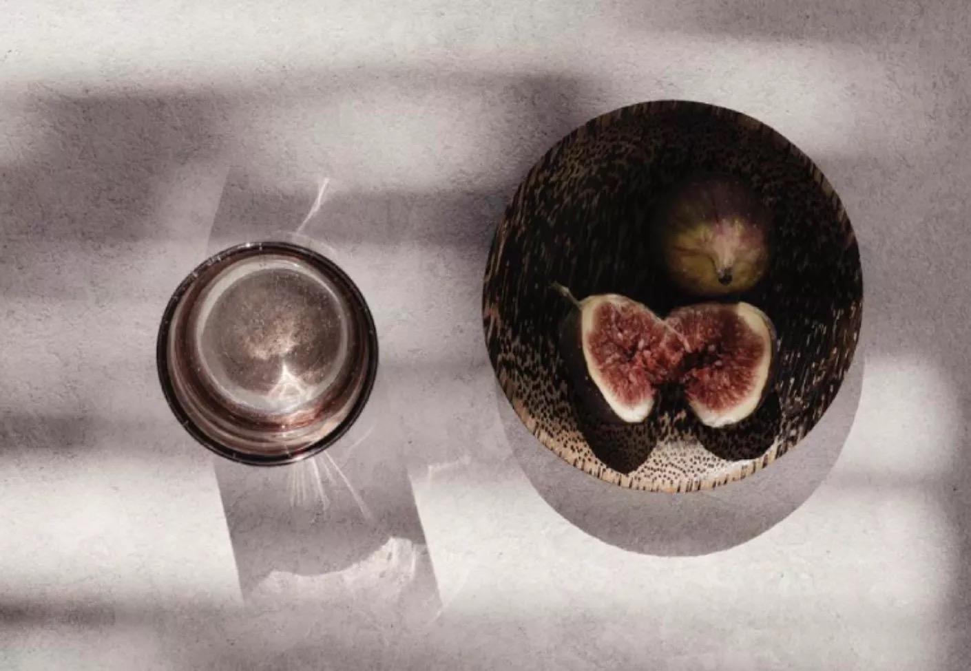 A small metal bowl and a wooden dish of fresh figs, one halved, set on a pale stone surface in striped light.