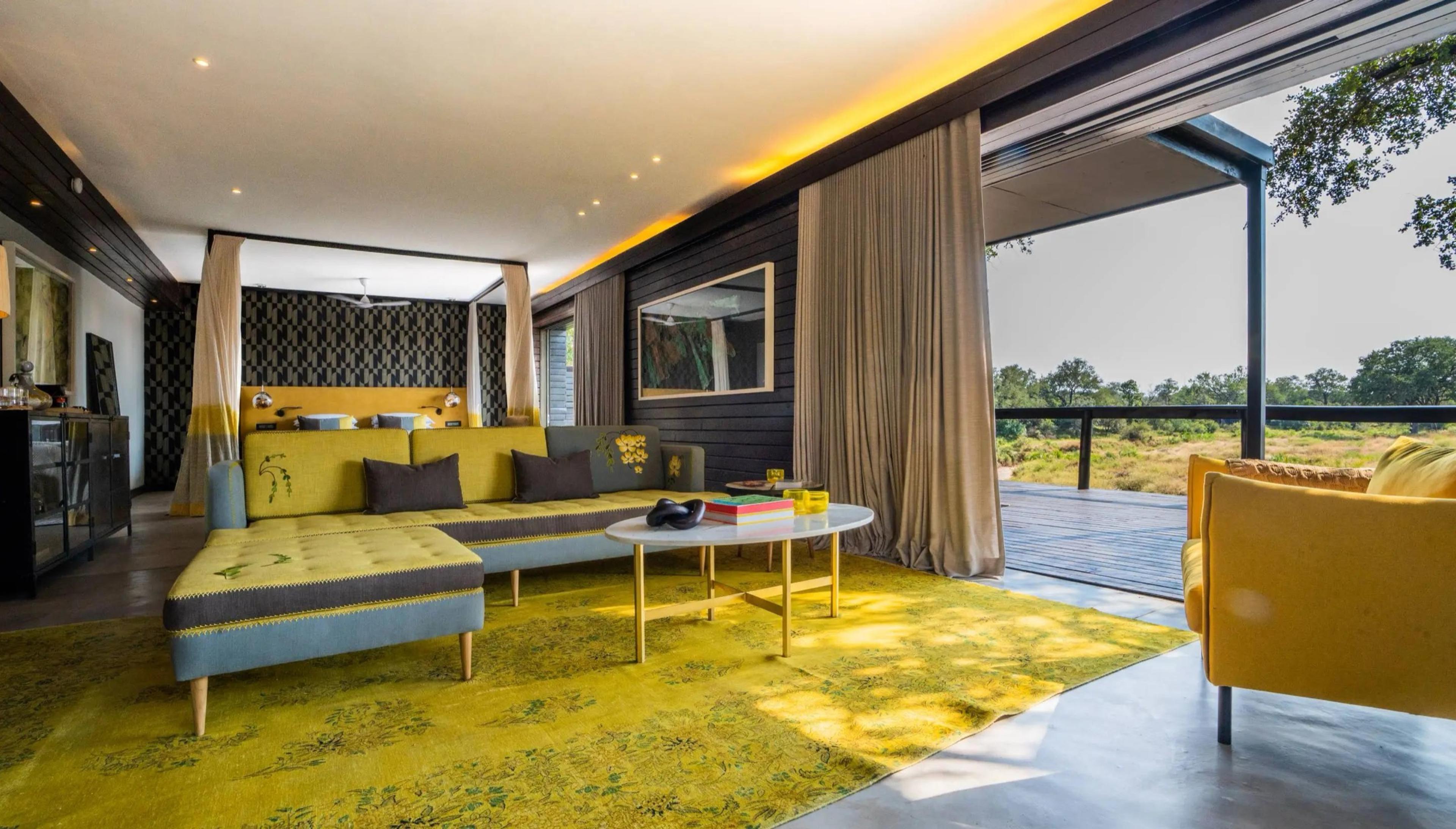 A lounge and bedroom area with a yellow sectional sofa and a yellow rug, next to sliding doors open onto a deck with trees beyond.