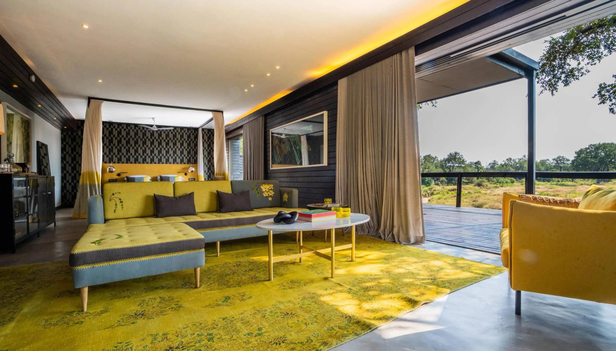 A lounge and bedroom area with a yellow sectional sofa and a yellow rug, next to sliding doors open onto a deck with trees beyond.